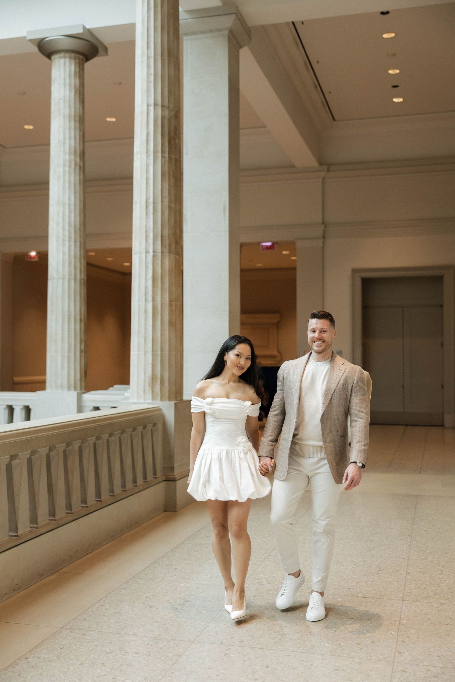 Elegant couple holding hands walking through grand museum hall with tall marble columns, woman in white mini dress, man in beige suit.