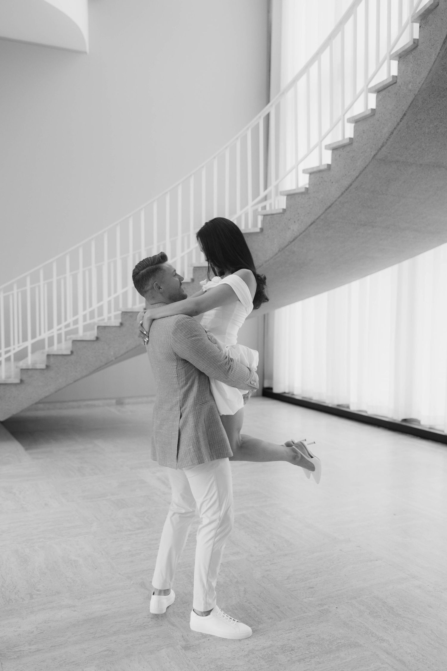 Black and white image of man lifting woman in white dress near spiral staircase in bright interior.