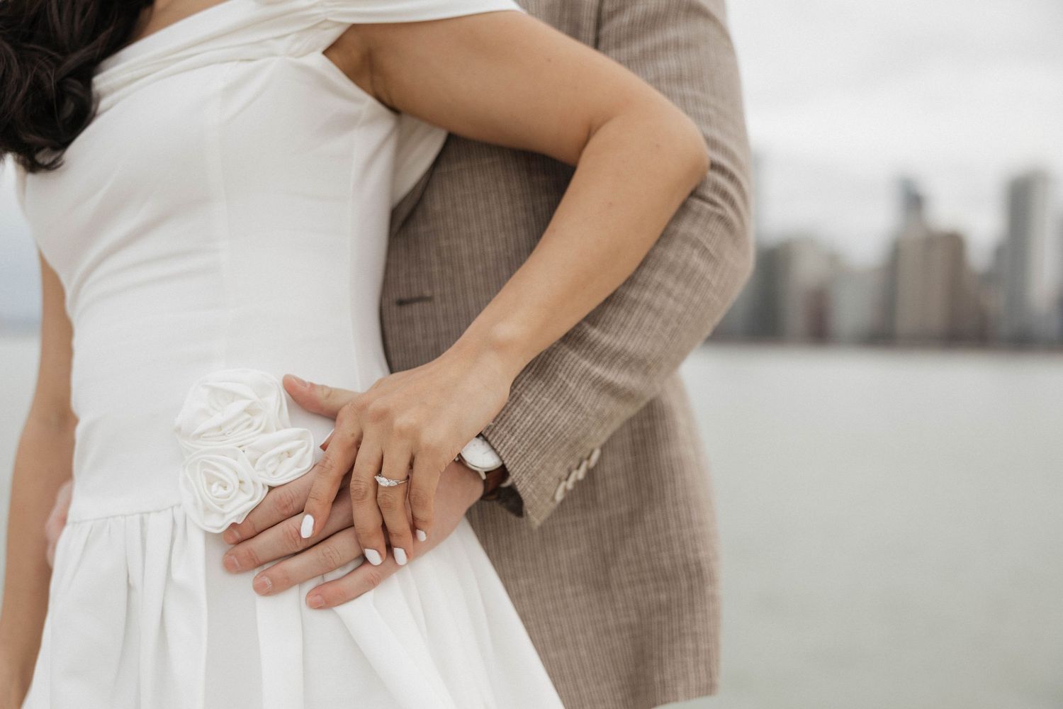 Couple embracing in wedding attire, showing off rings with city skyline in background.