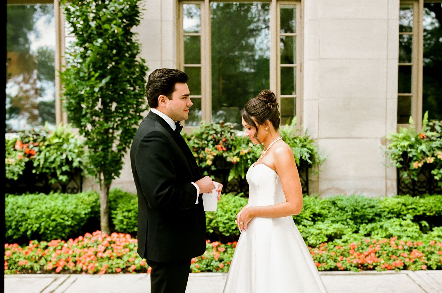 Bride and groom share an intimate first look moment outdoors, surrounded by lush green hedges and colorful flowers.