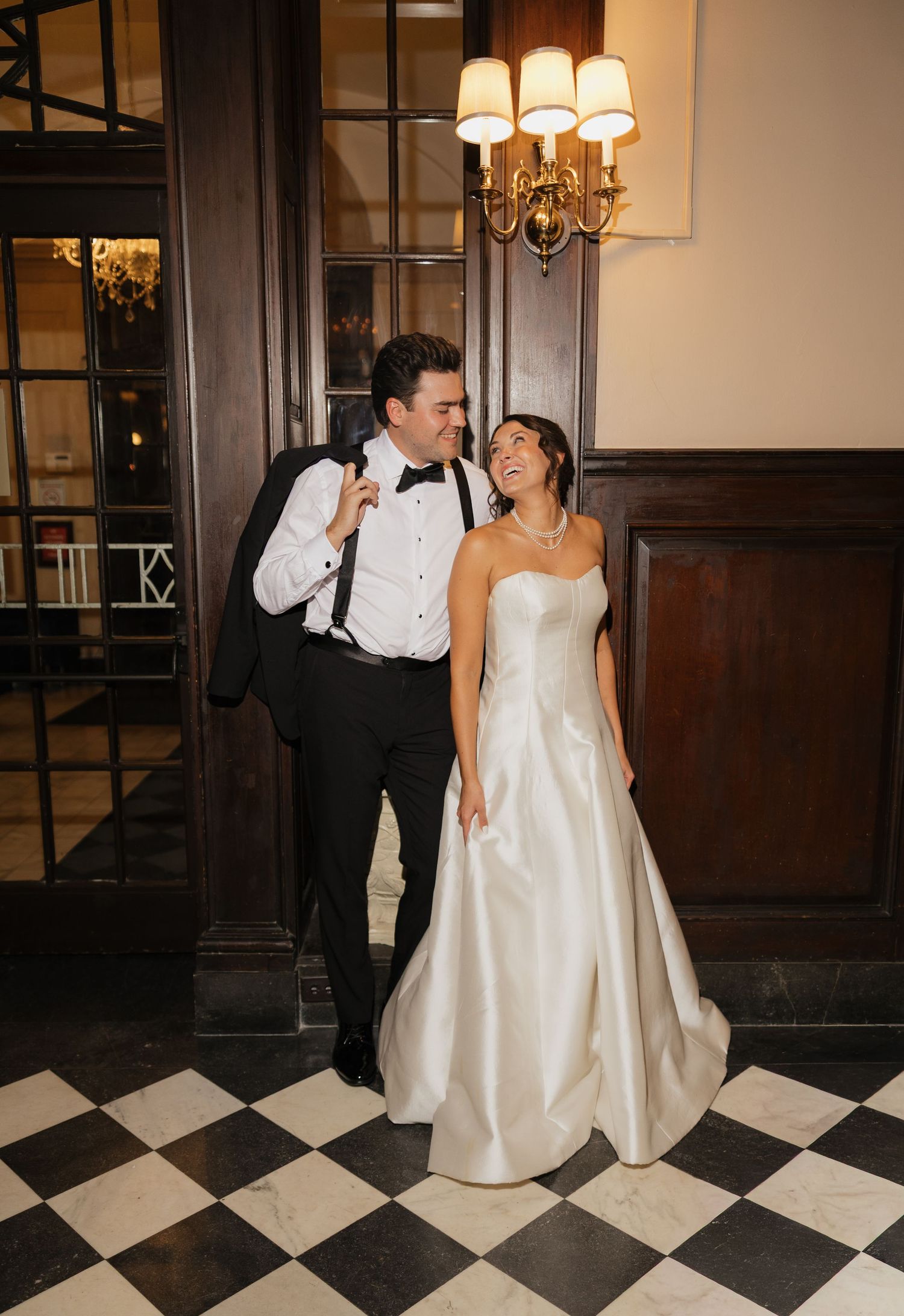 Bride in strapless satin gown and groom in black tuxedo share a joyful moment in an elegant venue with checkered floors.