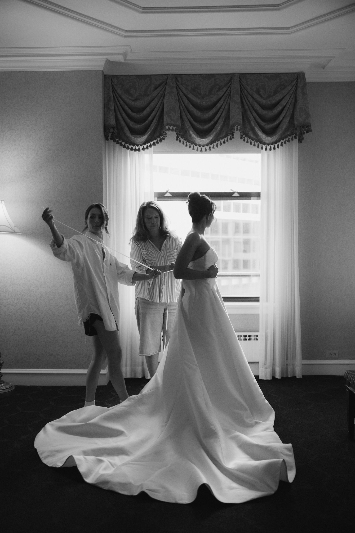Black and white photo of a bride in a flowing white gown being helped by two bridesmaids near a bright window.