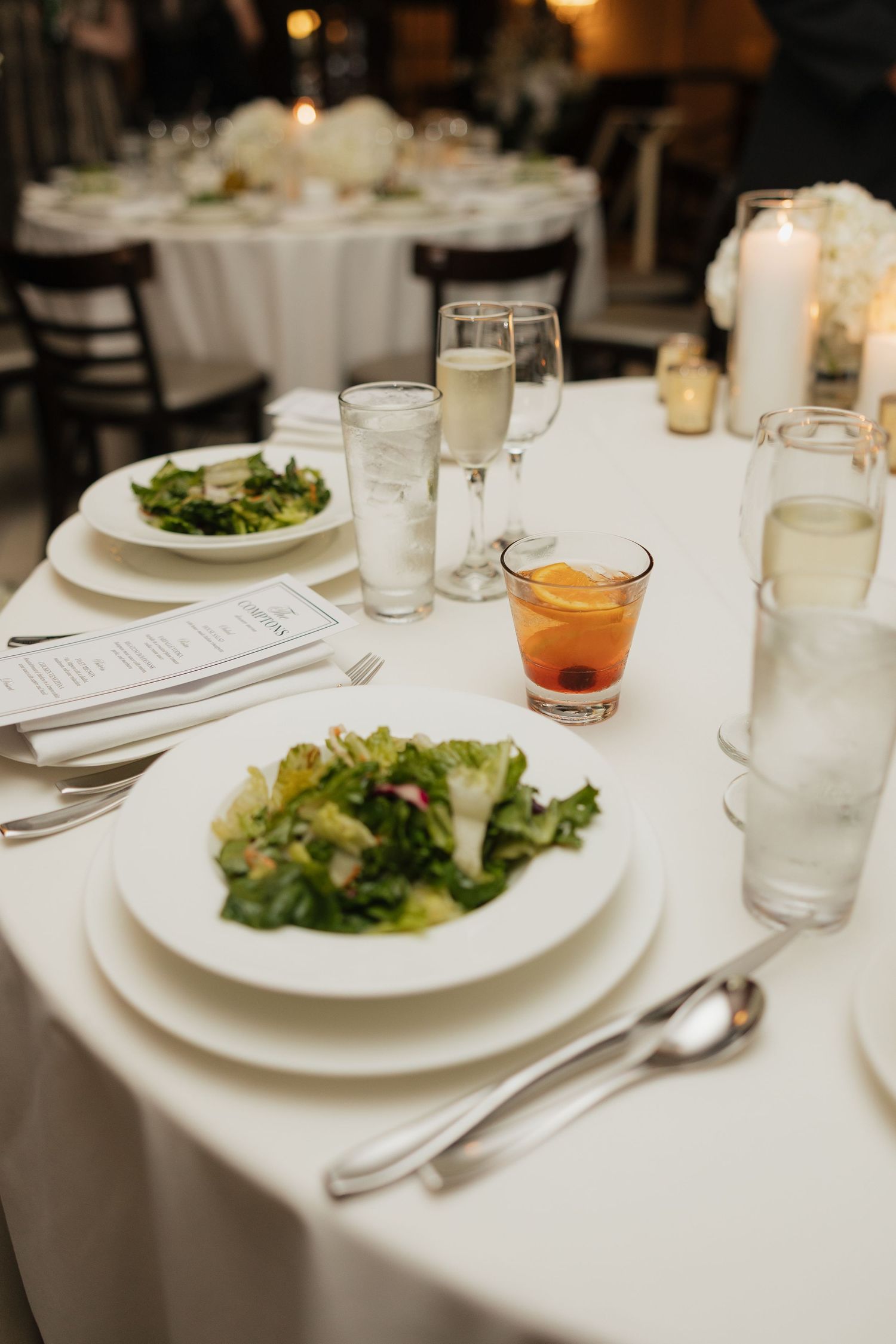 Elegant wedding reception table setting with fresh green salad, cocktail, champagne flute, and white linen tablecloth.