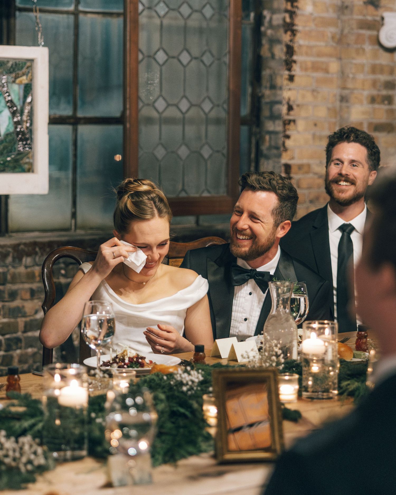 Wedding guests laugh and smile during an intimate candlelit dinner reception at a rustic brick venue.