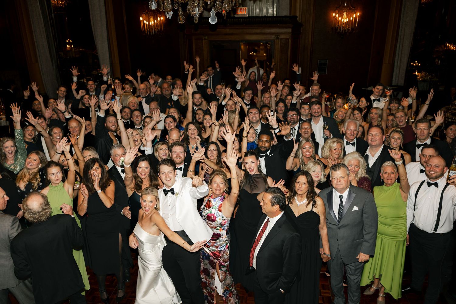 Large wedding reception group photo with bride and groom surrounded by cheering guests in elegant ballroom.