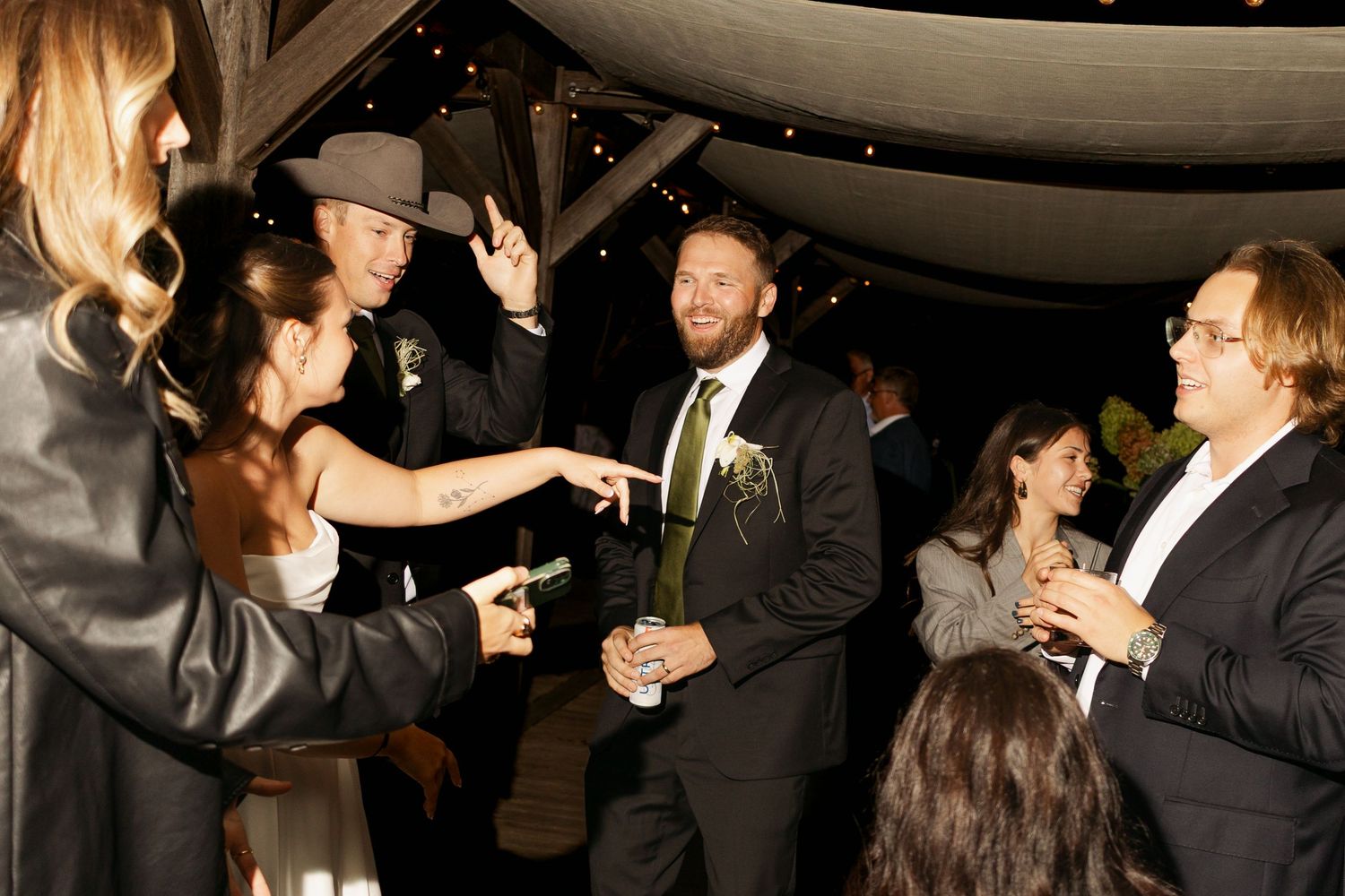 Wedding guests in formal attire laugh and mingle at an evening reception under a tent, with a bearded man in a dark suit and boutonniere.