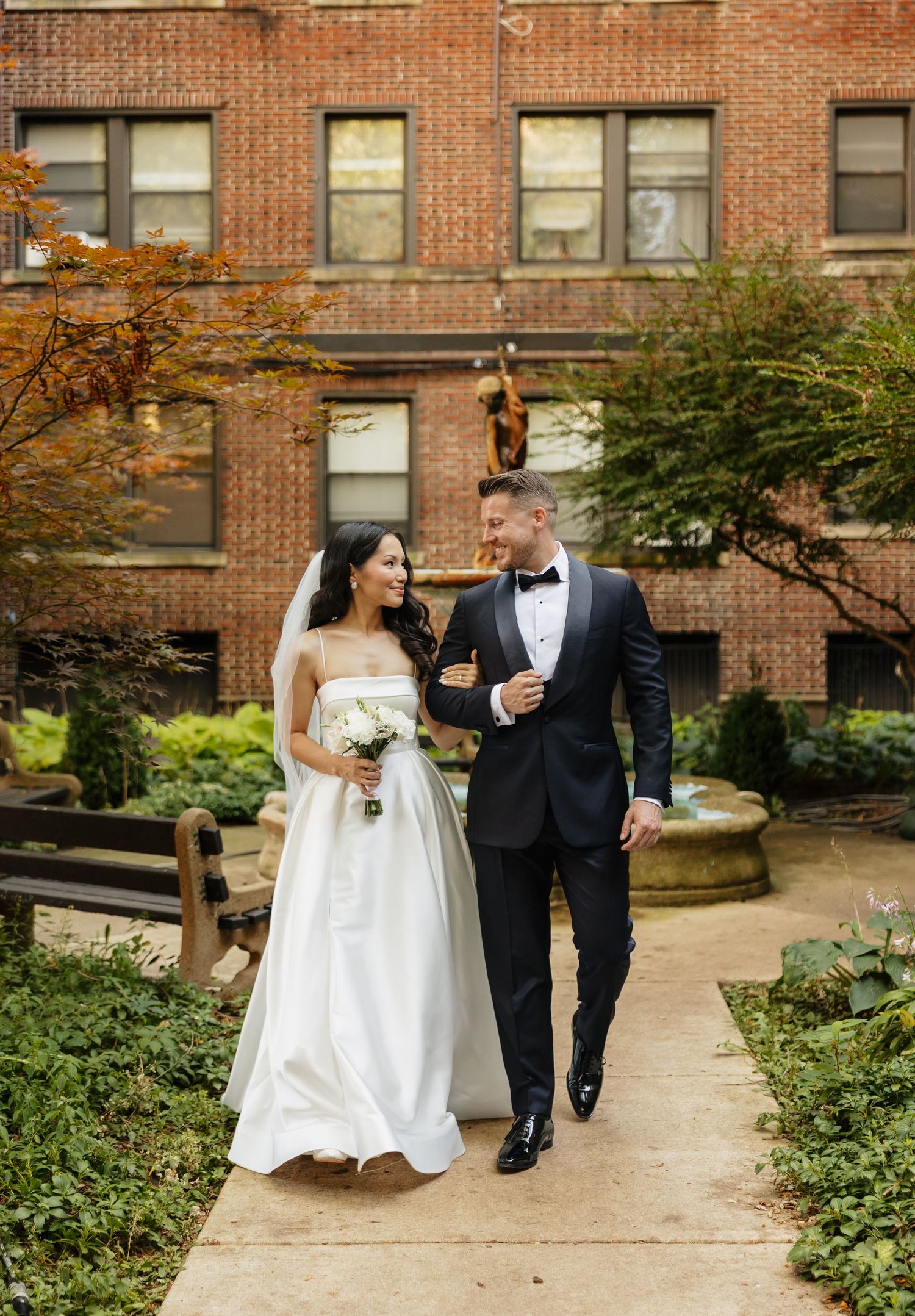 Bride in a white gown and groom in a black tuxedo walk arm in arm along a garden path in front of a red brick building.