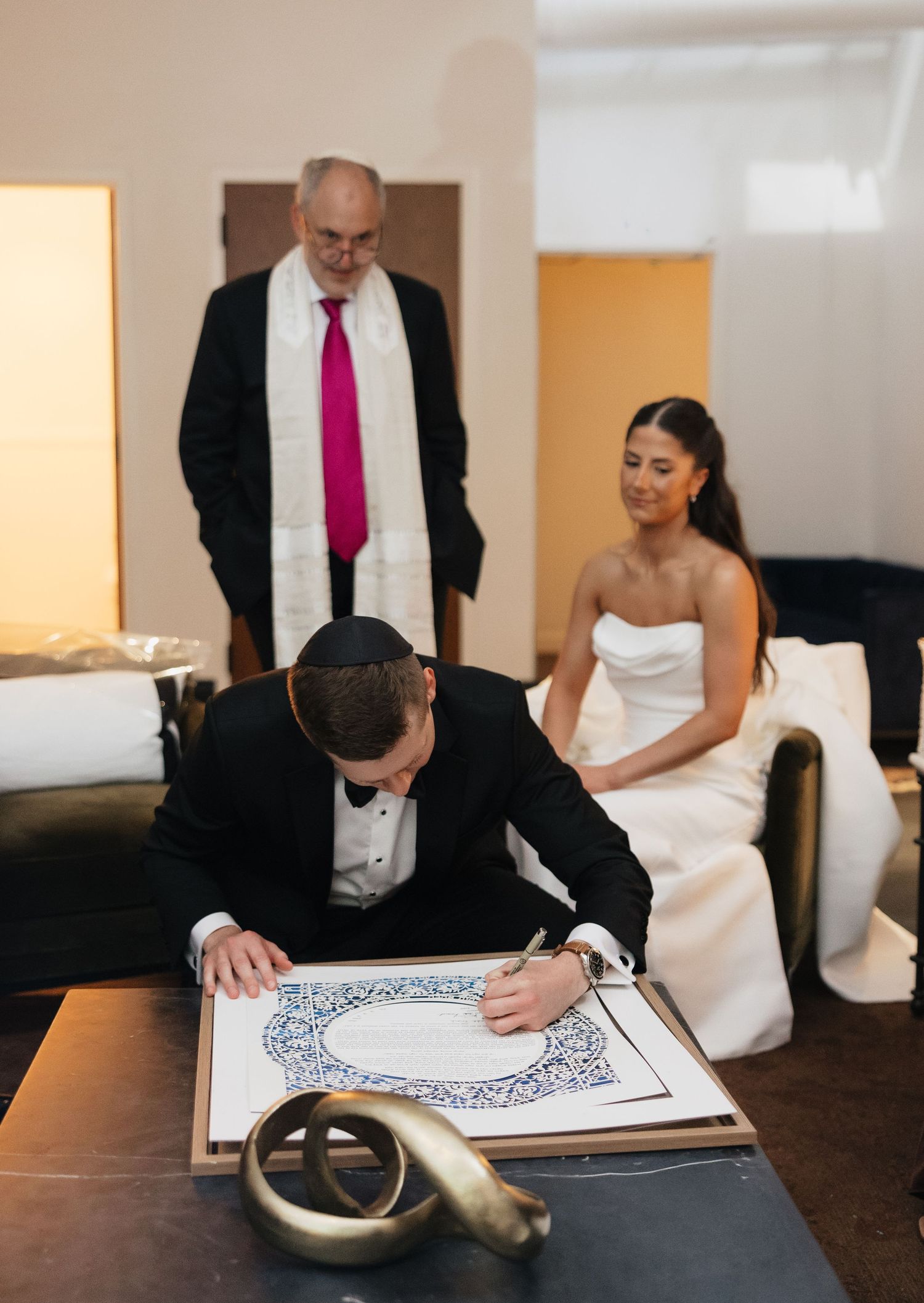 Groom signing Jewish ketubah marriage contract while bride watches and officiant stands behind during wedding ceremony.