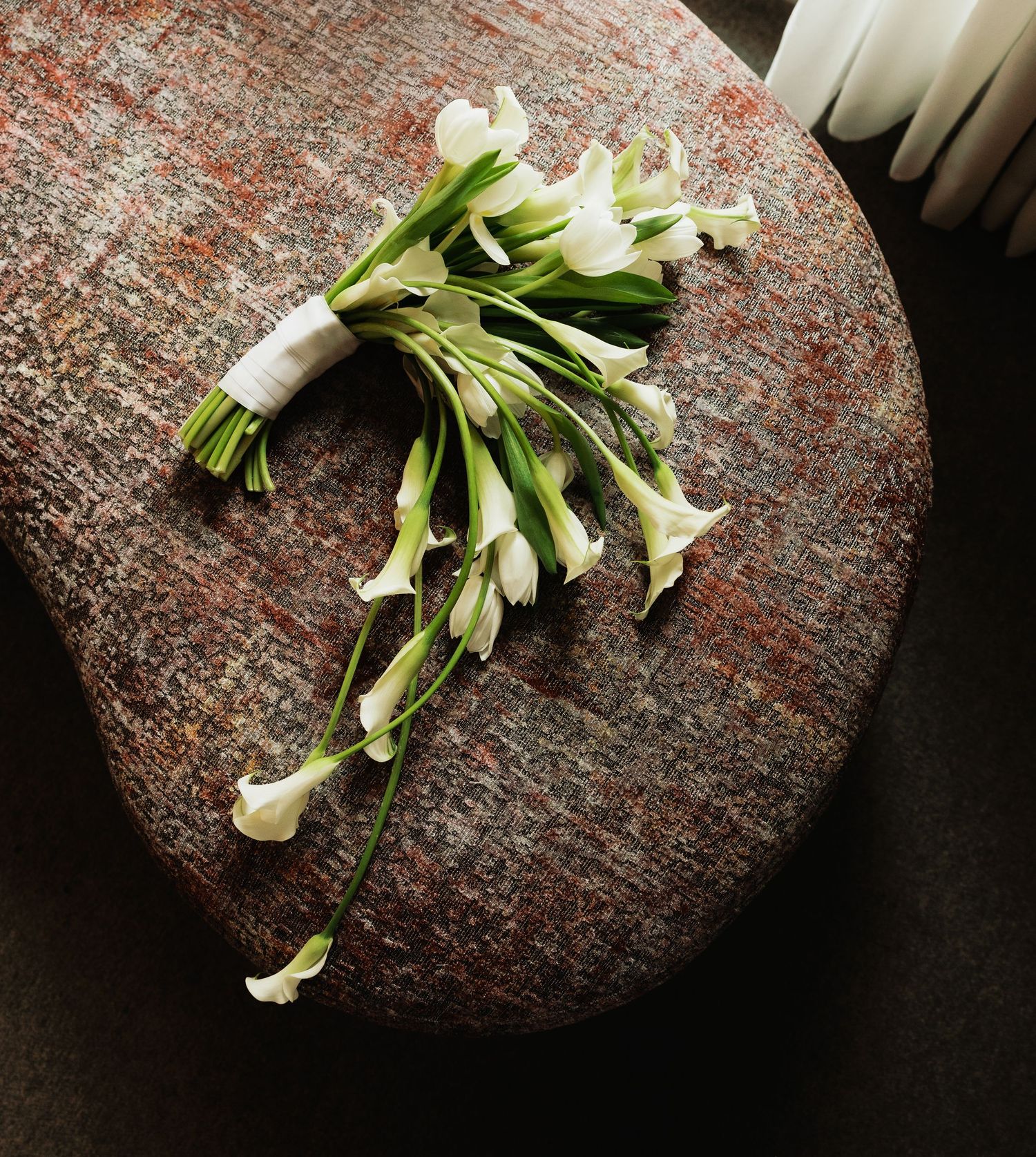 Elegant white calla lily wedding bouquet with green stems tied with white ribbon, resting on a rustic stone surface.