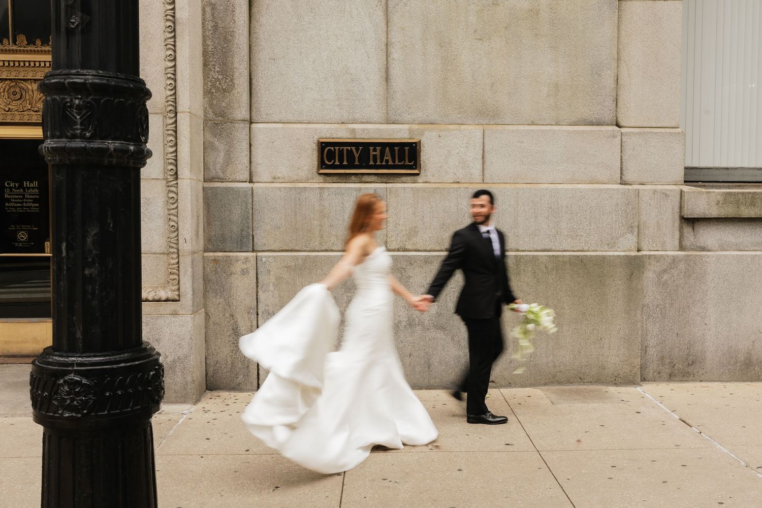 A bride and groom walk hand-in-hand past a City Hall building, her white gown flowing in motion.