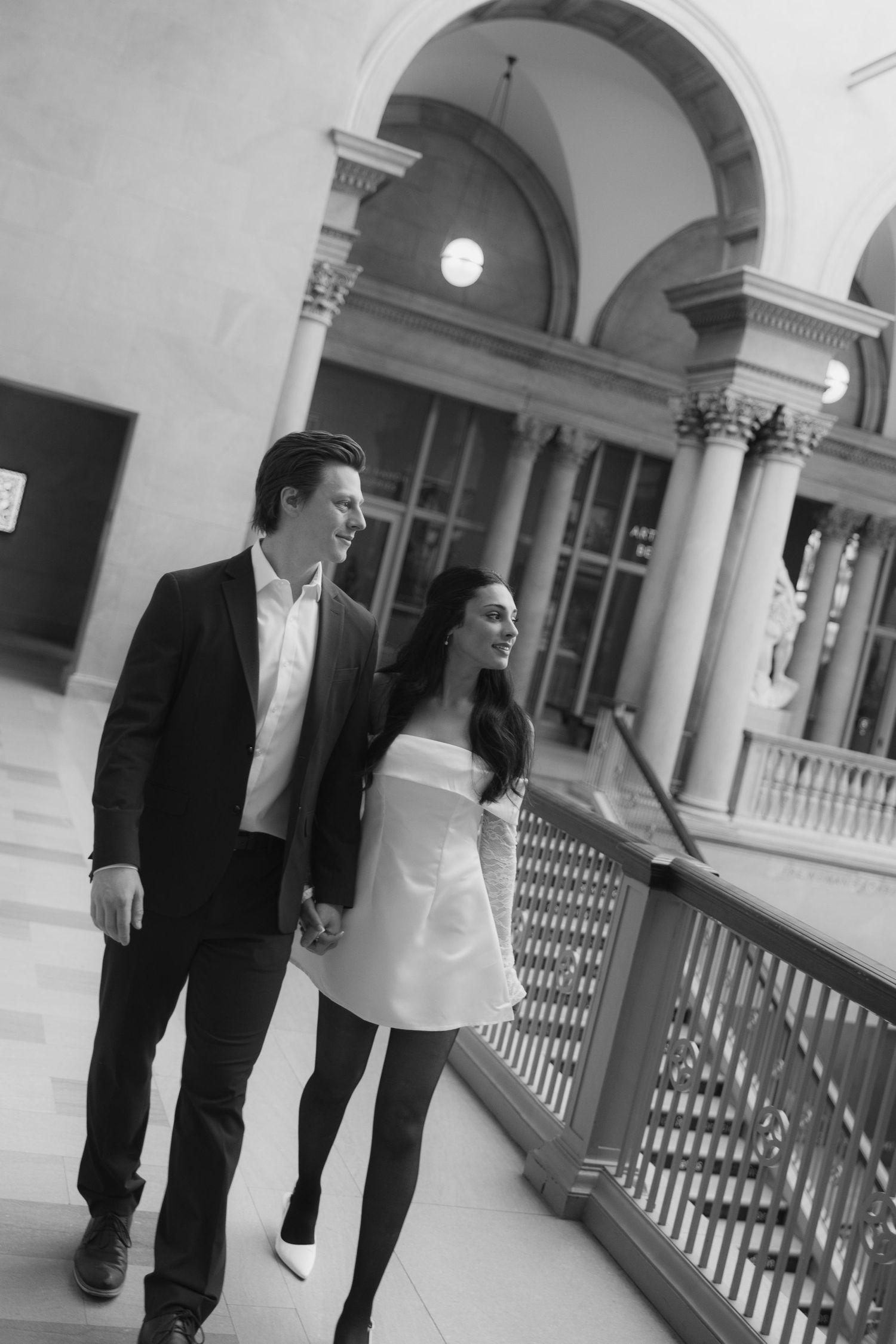 Two people walking through an ornate hallway with arched ceilings and columns.