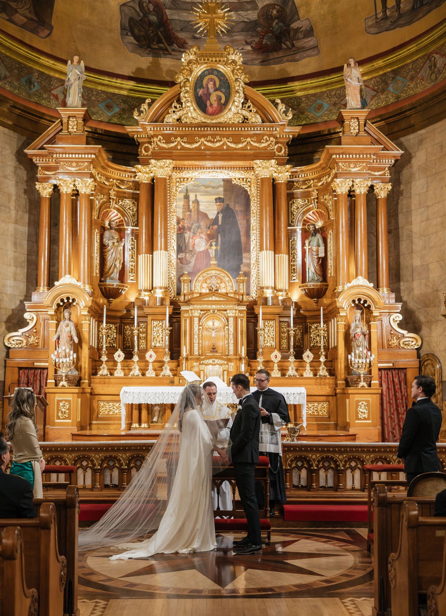 A couple stands at the altar of an ornate baroque church with golden decorative elements and religious artwork.
