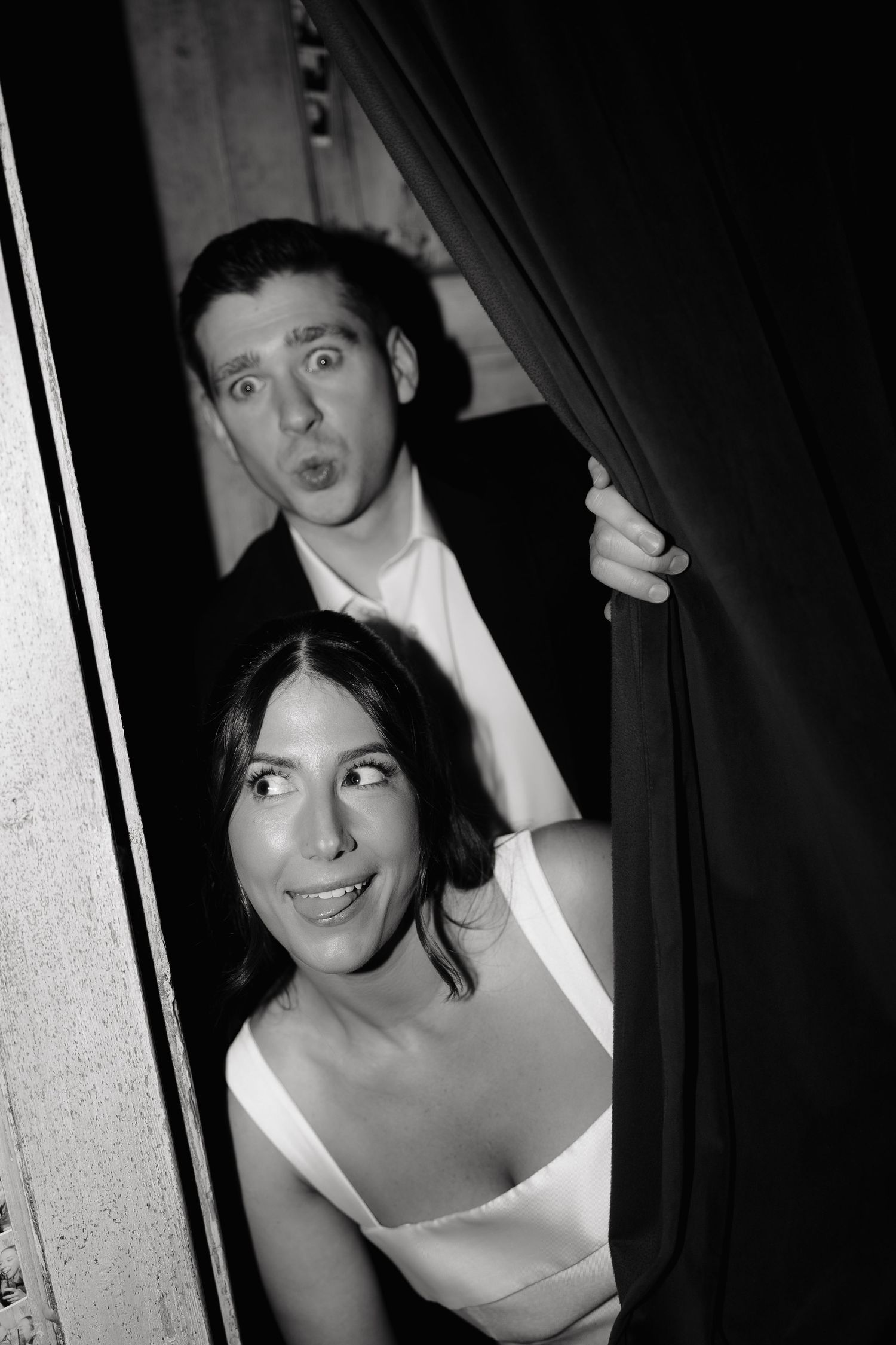 A black and white candid photo of a couple peeking playfully through curtains.