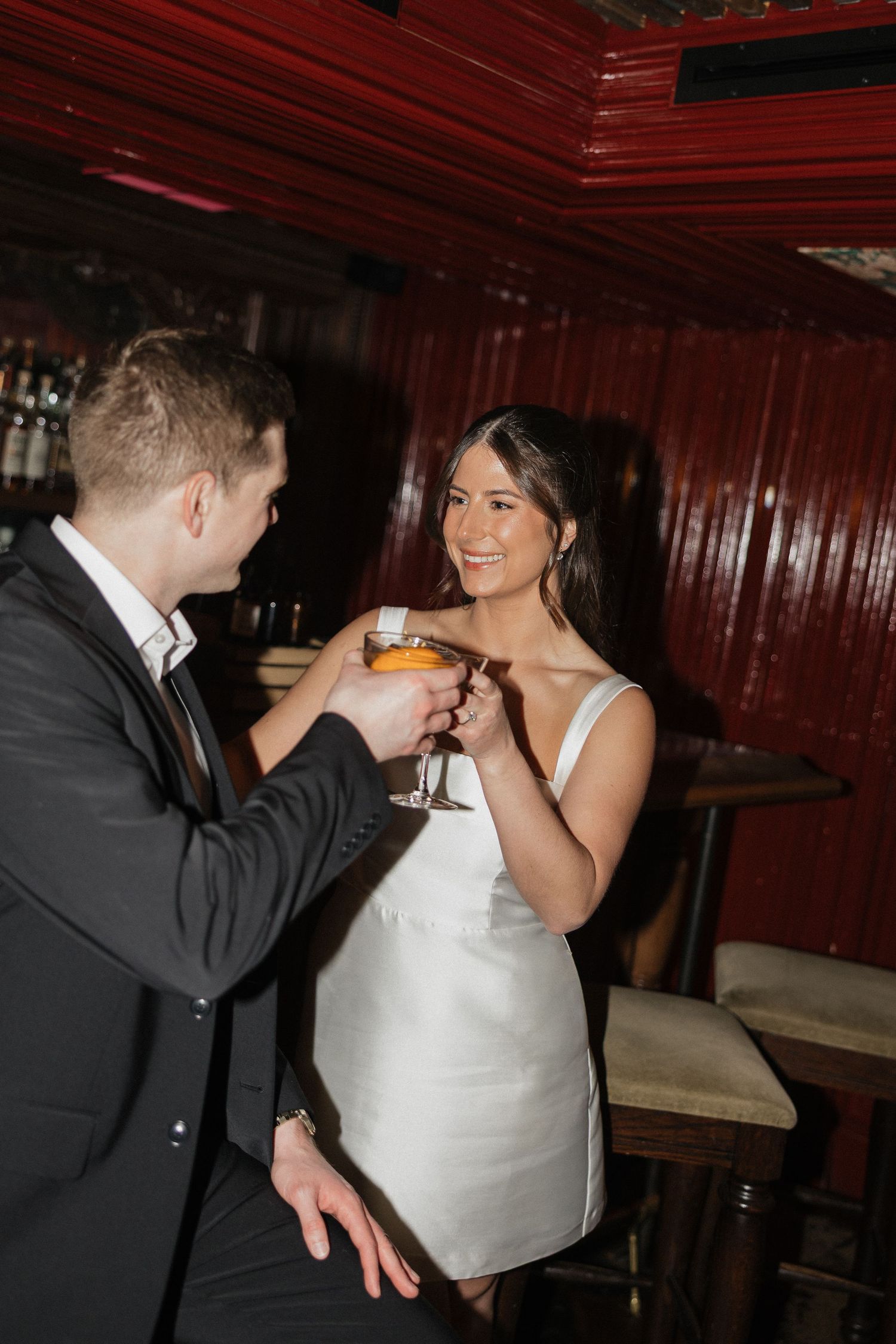 A couple in formal wedding attire shares a romantic toast at a dimly lit venue with dark wood paneling.