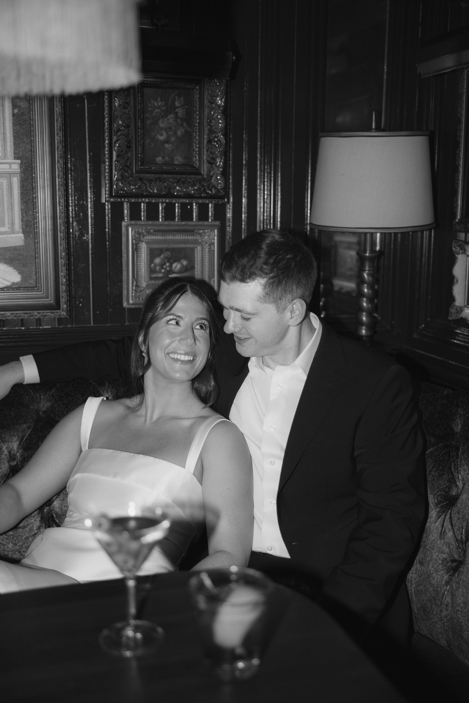 A couple in formal attire share an intimate moment at a dimly lit bar with cocktails and vintage decor.