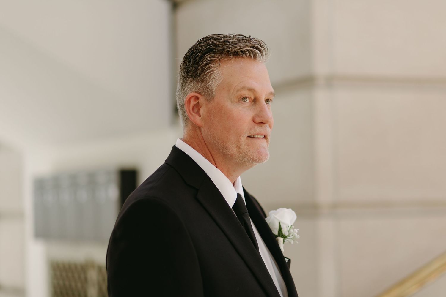 A person in a black suit and white boutonniere stands in an elegant indoor setting.