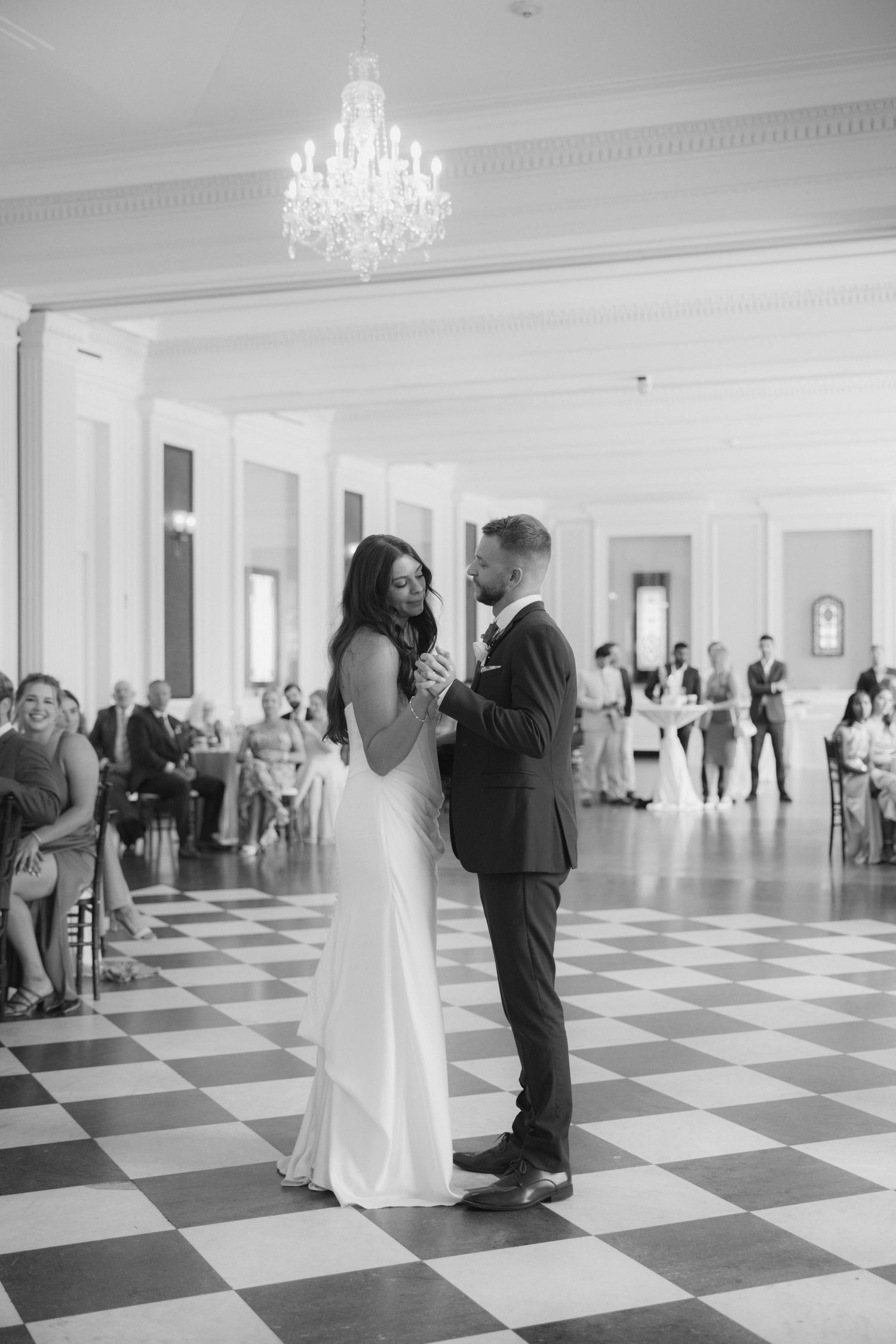 A couple shares their first dance in an elegant ballroom with a black and white checkered floor and crystal chandeliers.