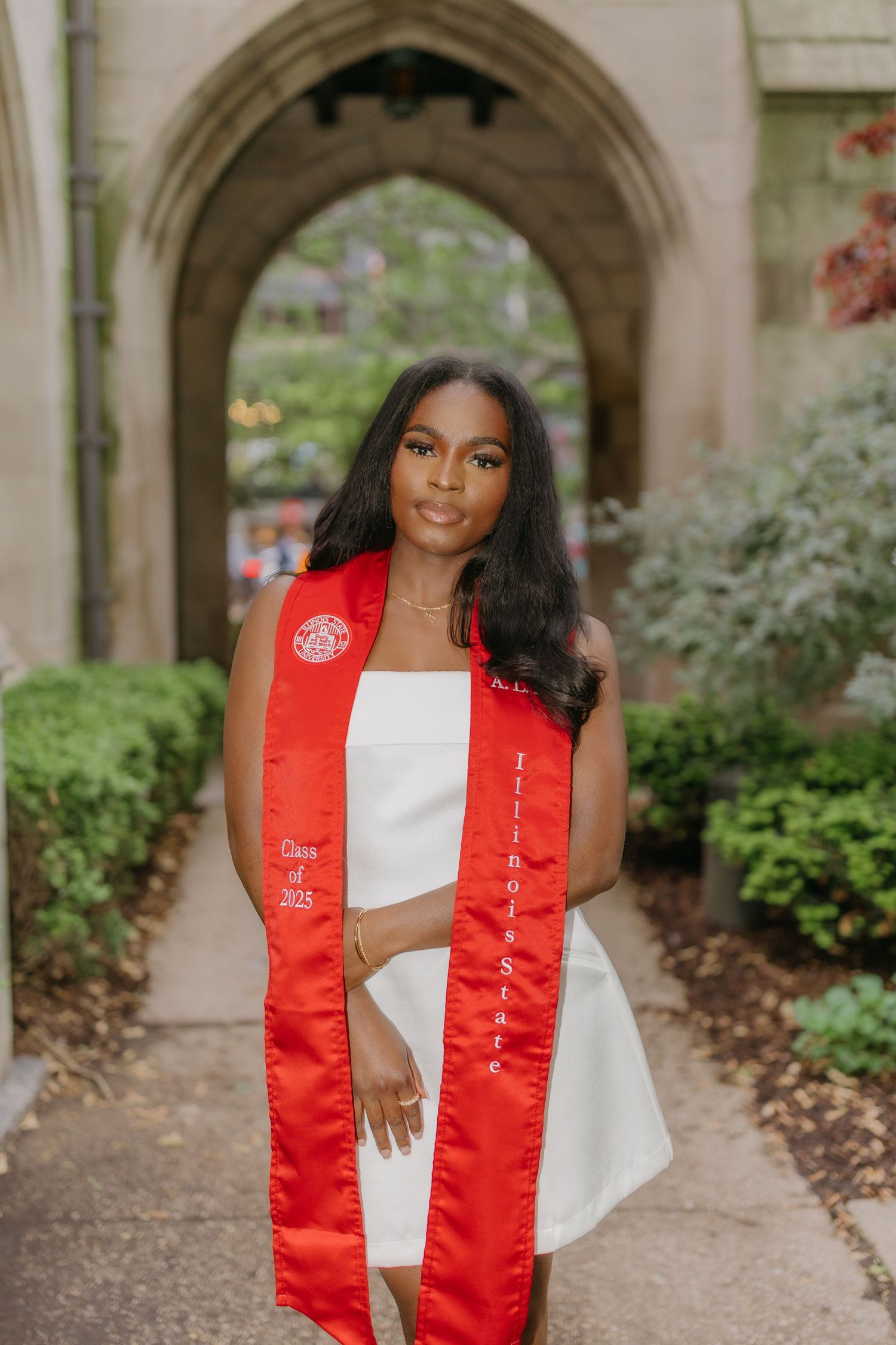 Graduate in white dress and red graduation stole poses in outdoor archway for professional portraits.