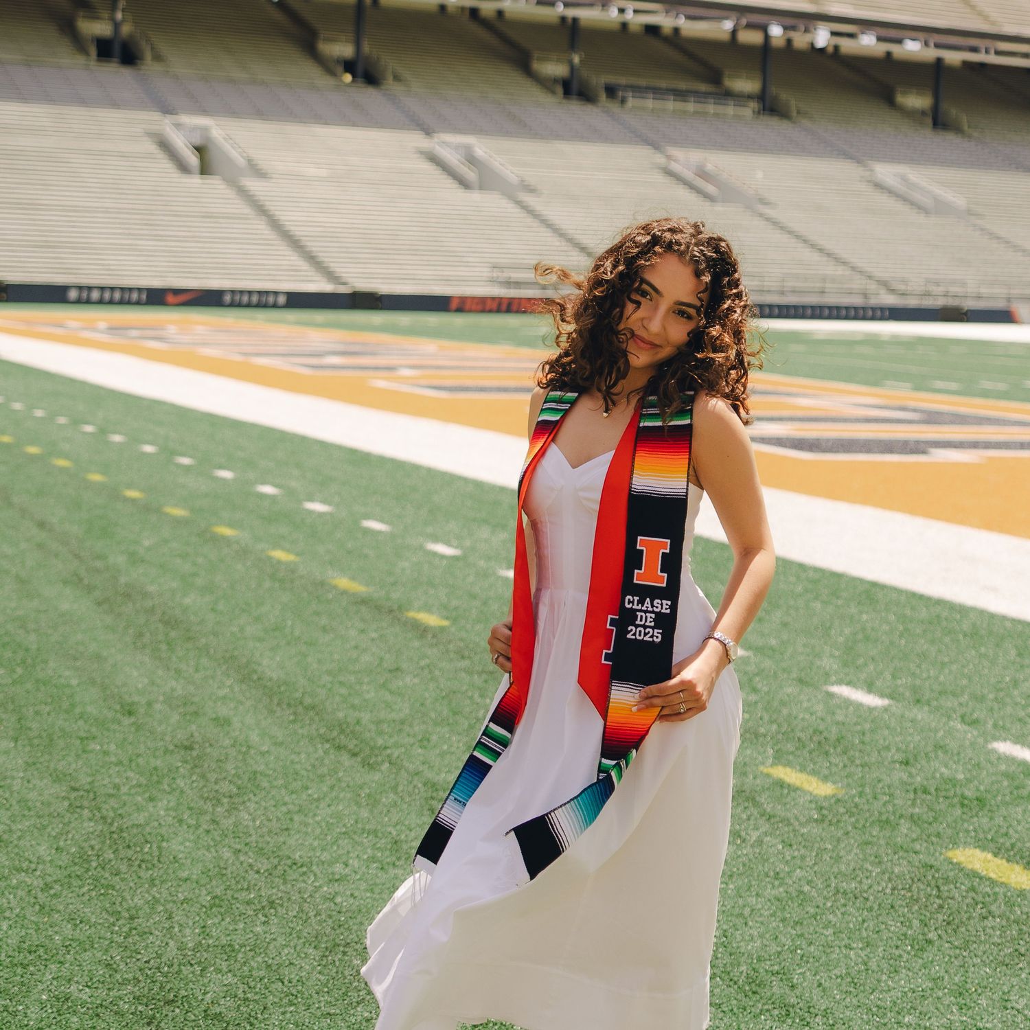 Graduate in white dress and red graduation stole poses on football field during commencement photoshoot.