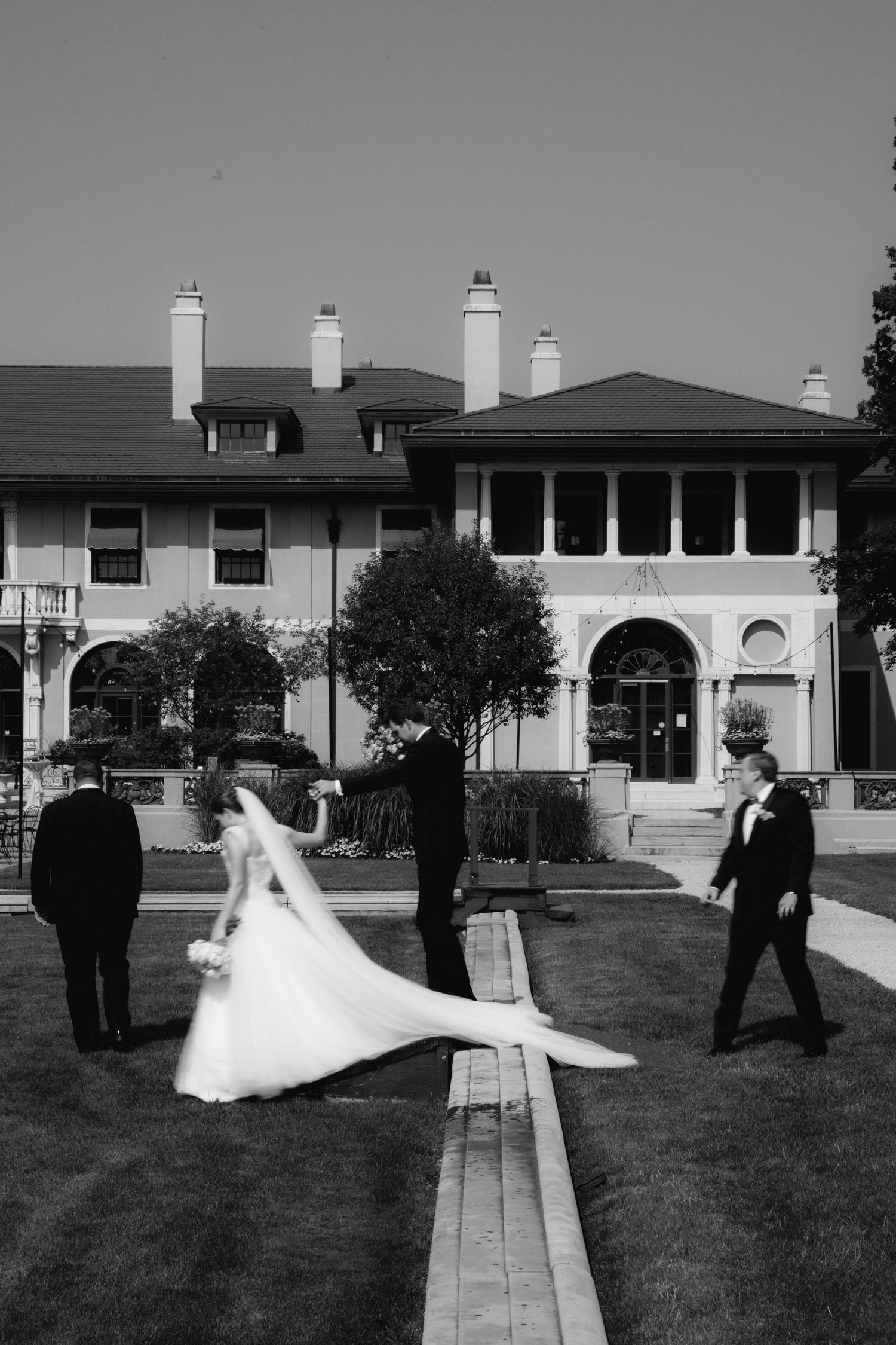Black and white photo of bride in flowing gown with veil and groomsmen outside grand estate venue.