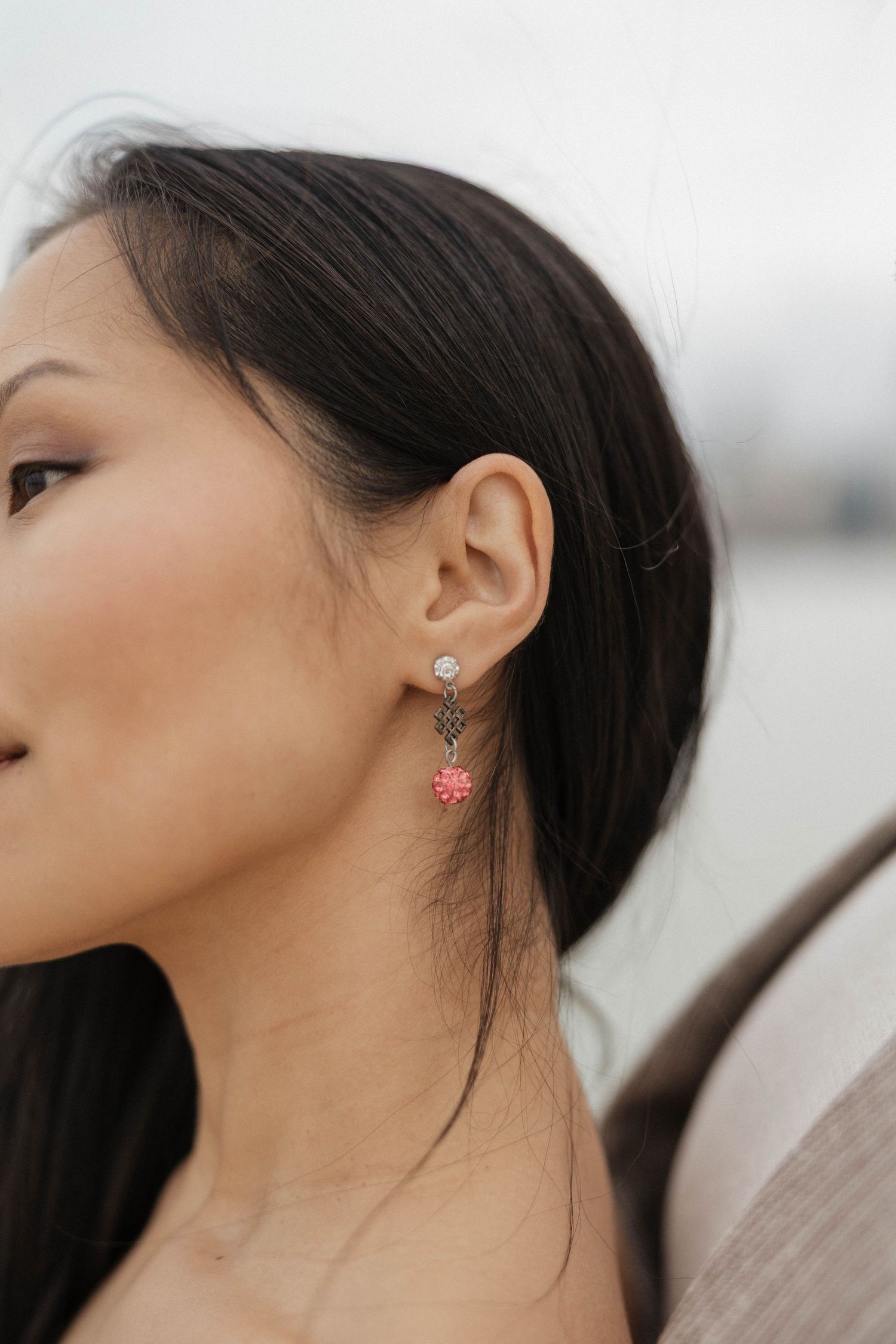 Woman wearing elegant drop earrings with crystal stud, dark chain, and pink floral charm against city backdrop.