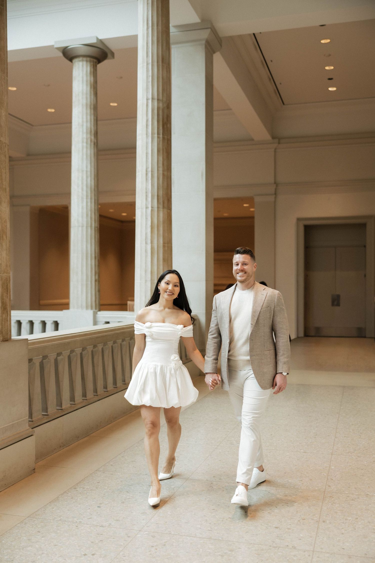 Happy couple walking together in a grand neoclassical building, woman wearing white off-shoulder dress, man in tan blazer.