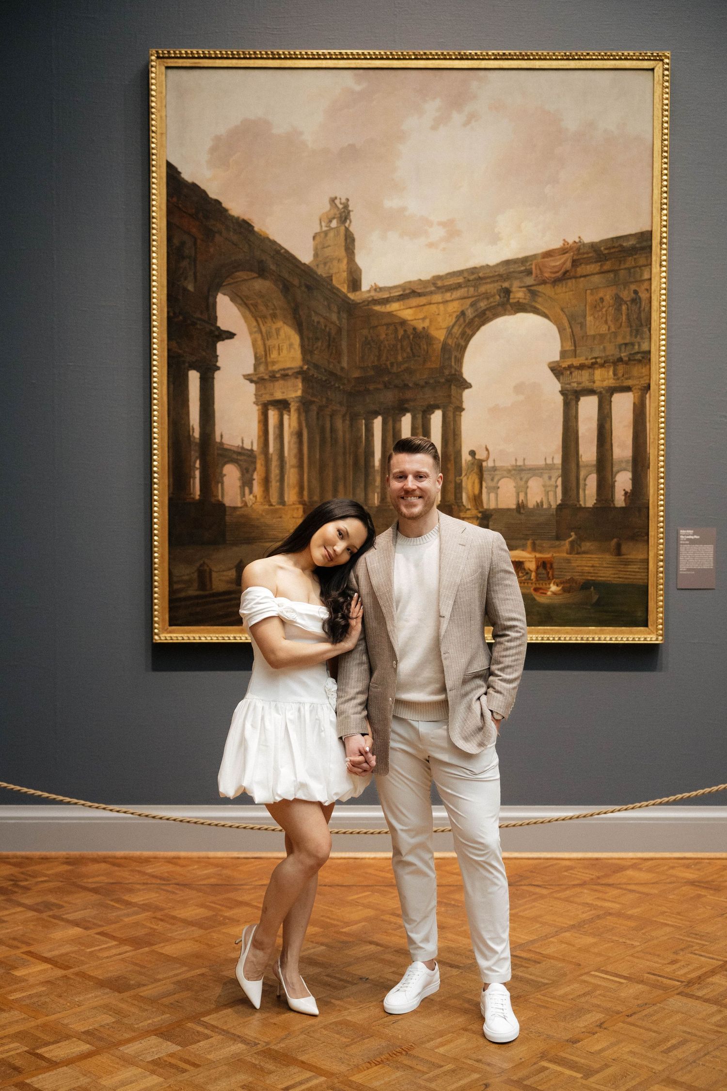 Couple posing hand-in-hand in white and beige outfits before a classical ruins painting in an art museum.
