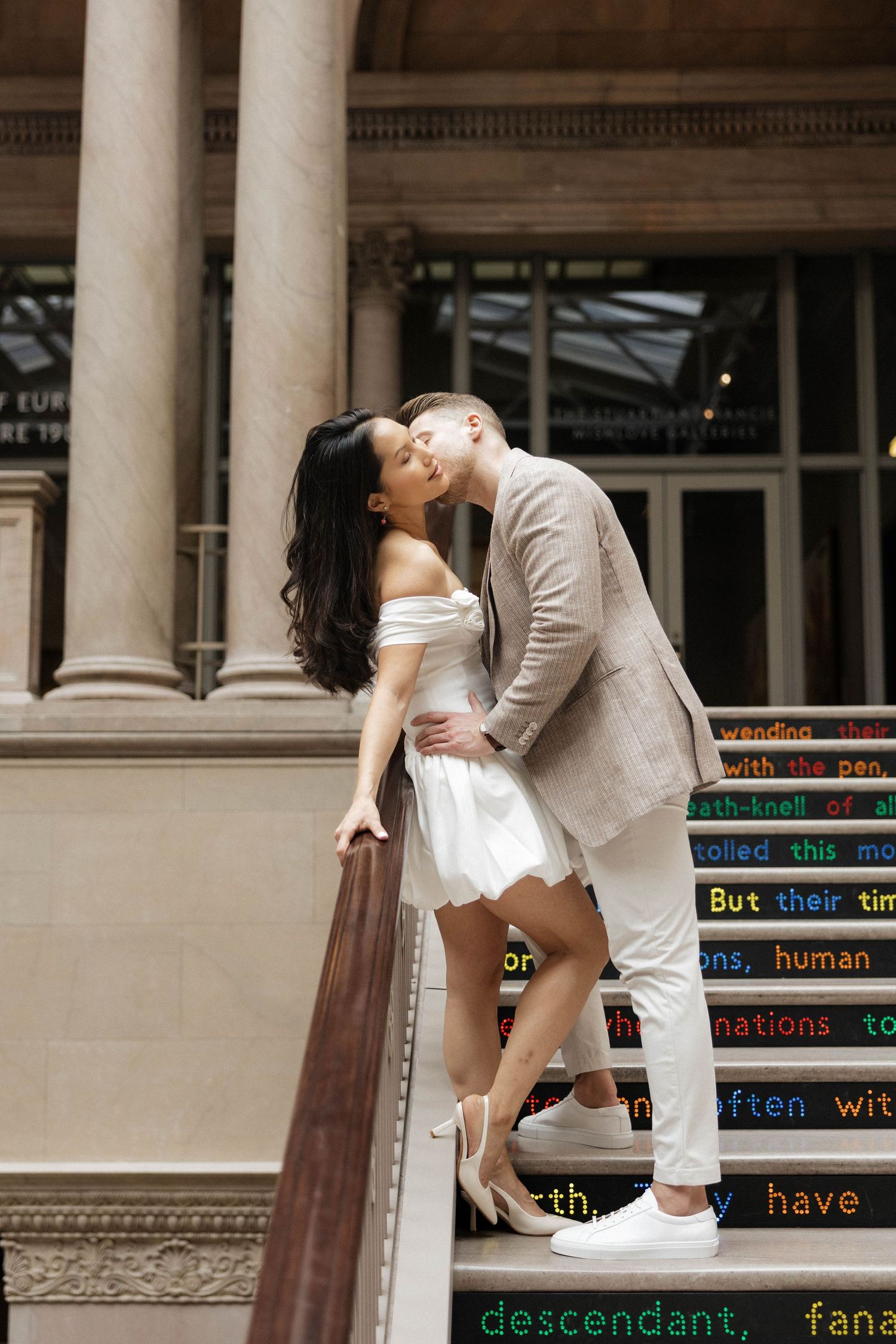 Couple sharing a romantic kiss on ornate building steps, woman in white mini dress and man in beige suit.