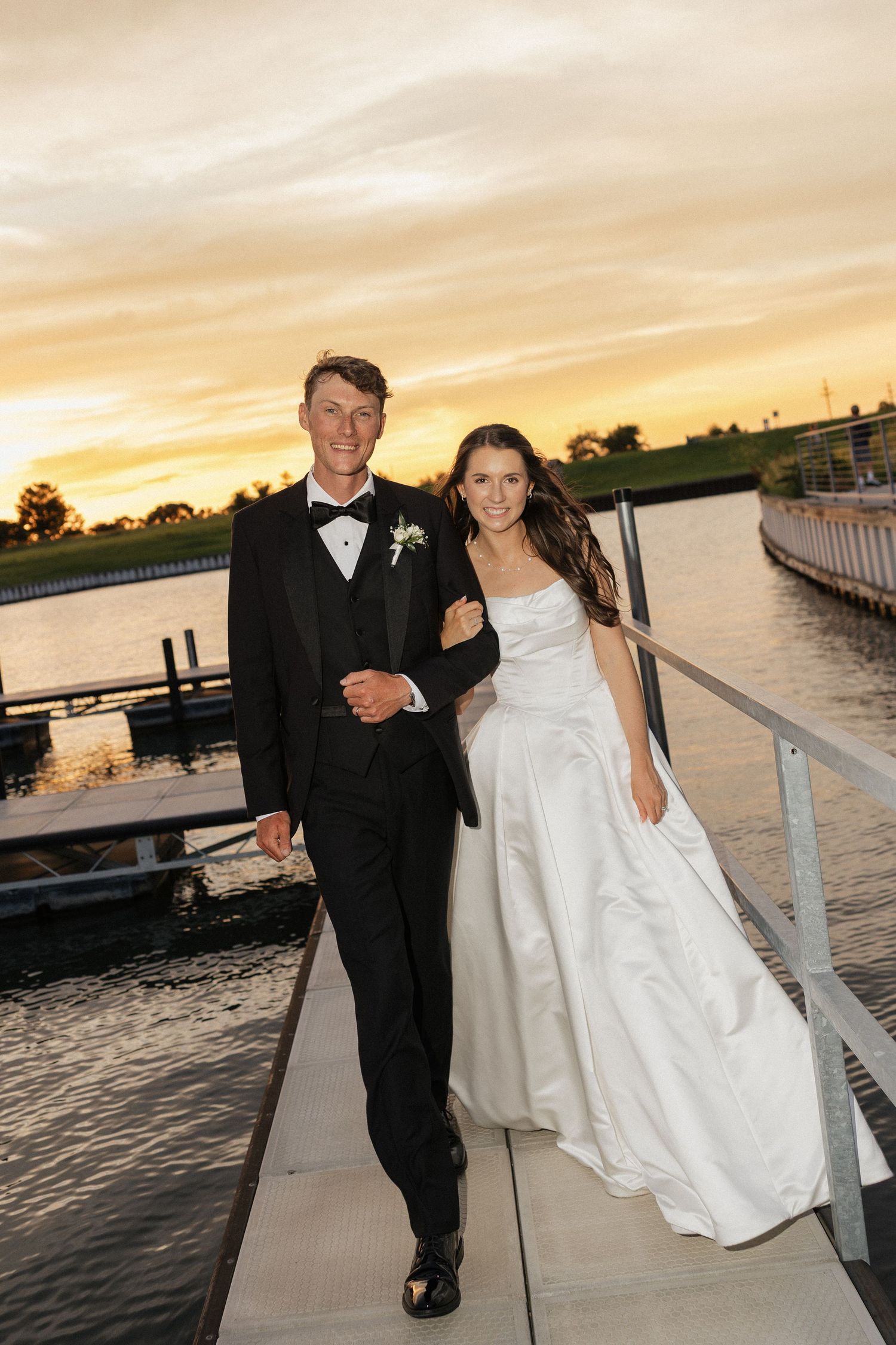 Bride and groom walking on a waterfront dock at sunset, dressed in wedding attire.