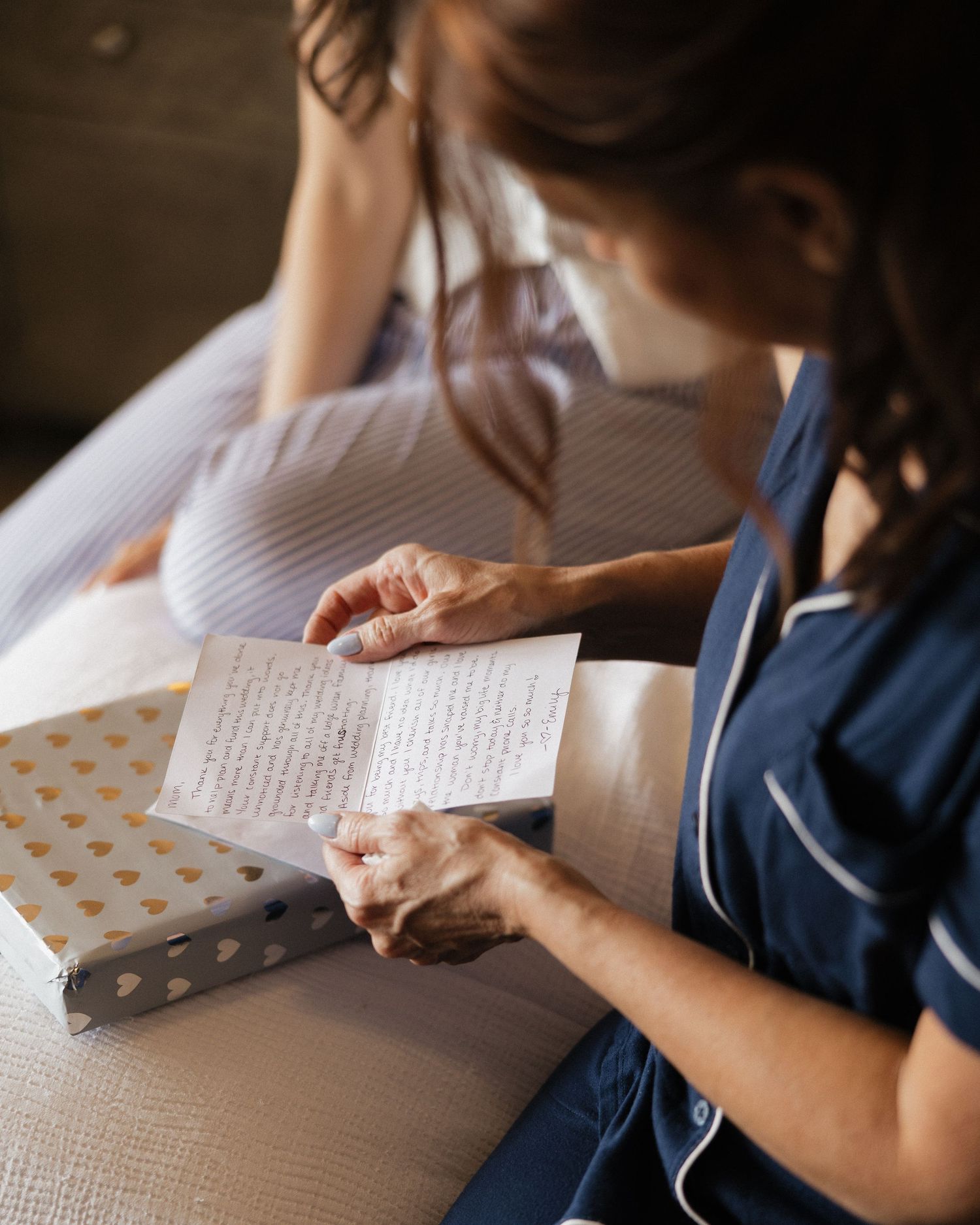 A nurse in navy scrubs reads a handwritten card next to a gift box with gold hearts beside a patient in bed.