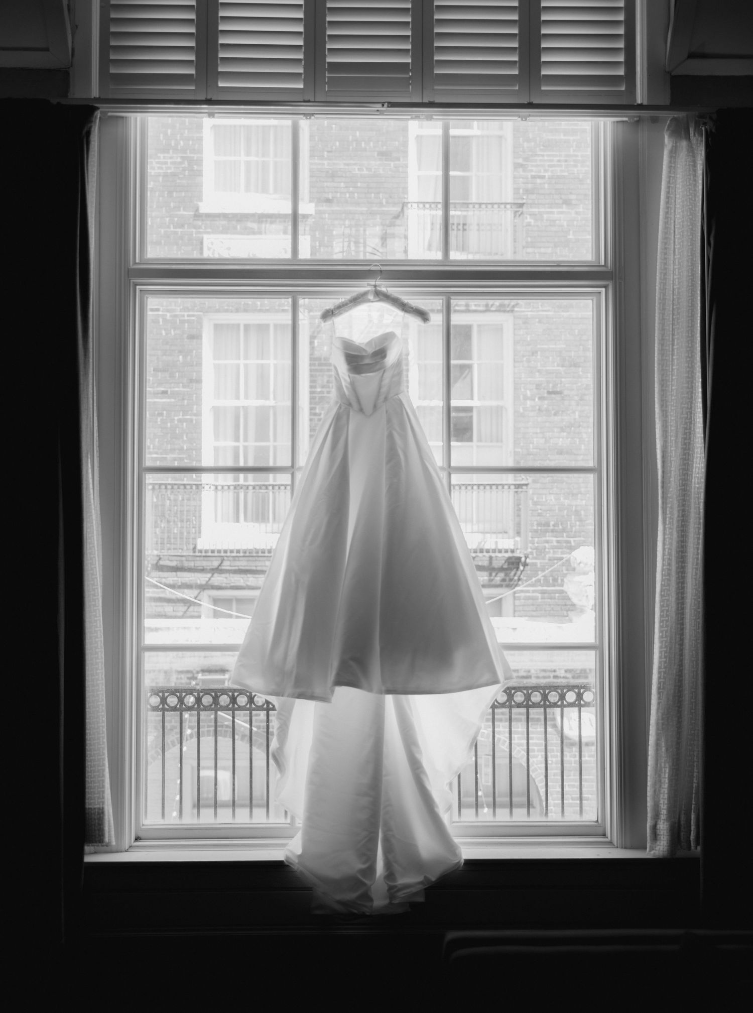 Black and white photo of an elegant strapless wedding gown hanging in a tall window with city buildings visible outside.