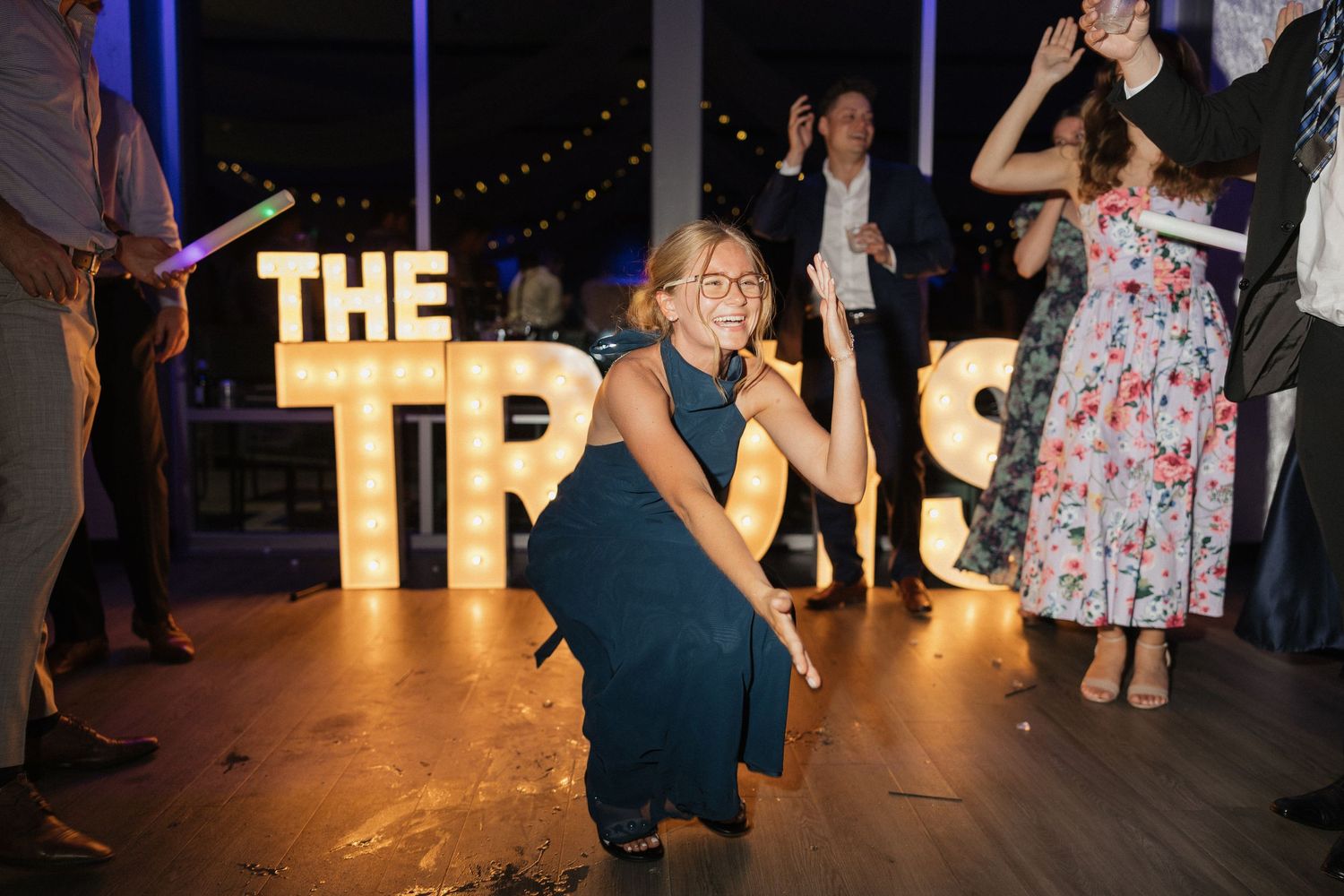 Woman in teal dress dancing at wedding reception near illuminated marquee letter sign reading The Tros.