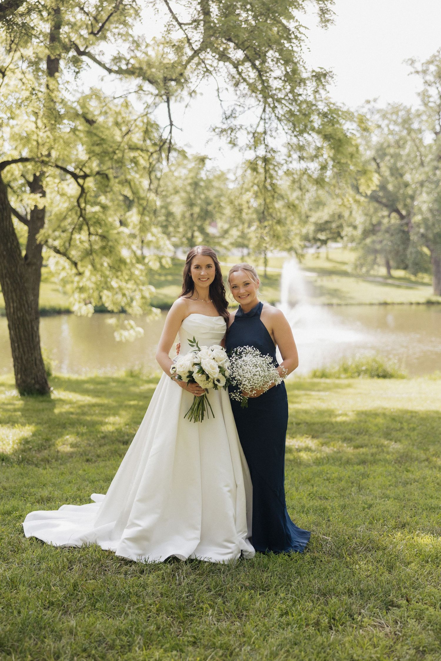 Bride in white gown and bridesmaid in navy dress holding white bouquets outdoors near a pond with fountain.