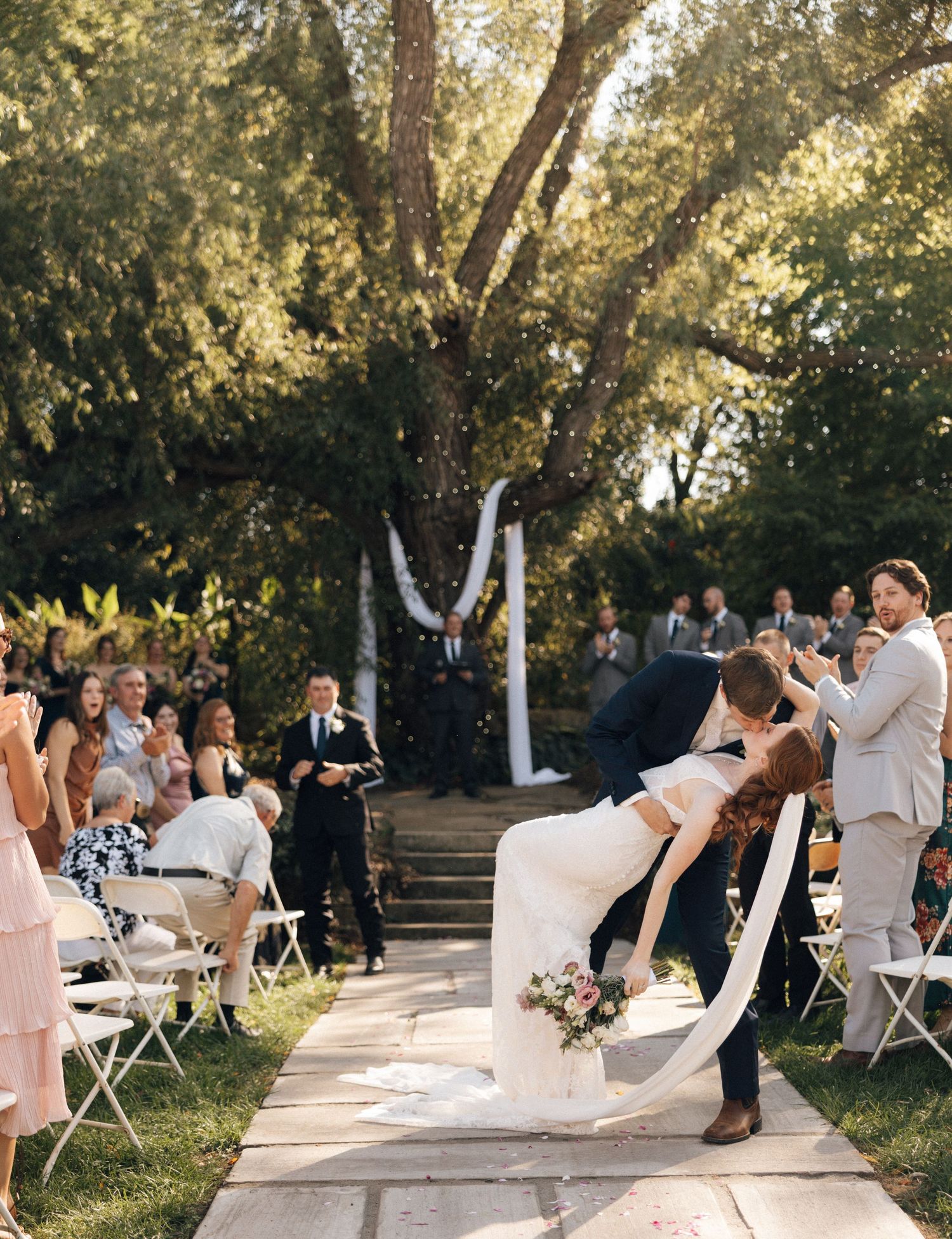 Groom dips bride in a romantic kiss during outdoor wedding ceremony aisle exit with guests cheering.