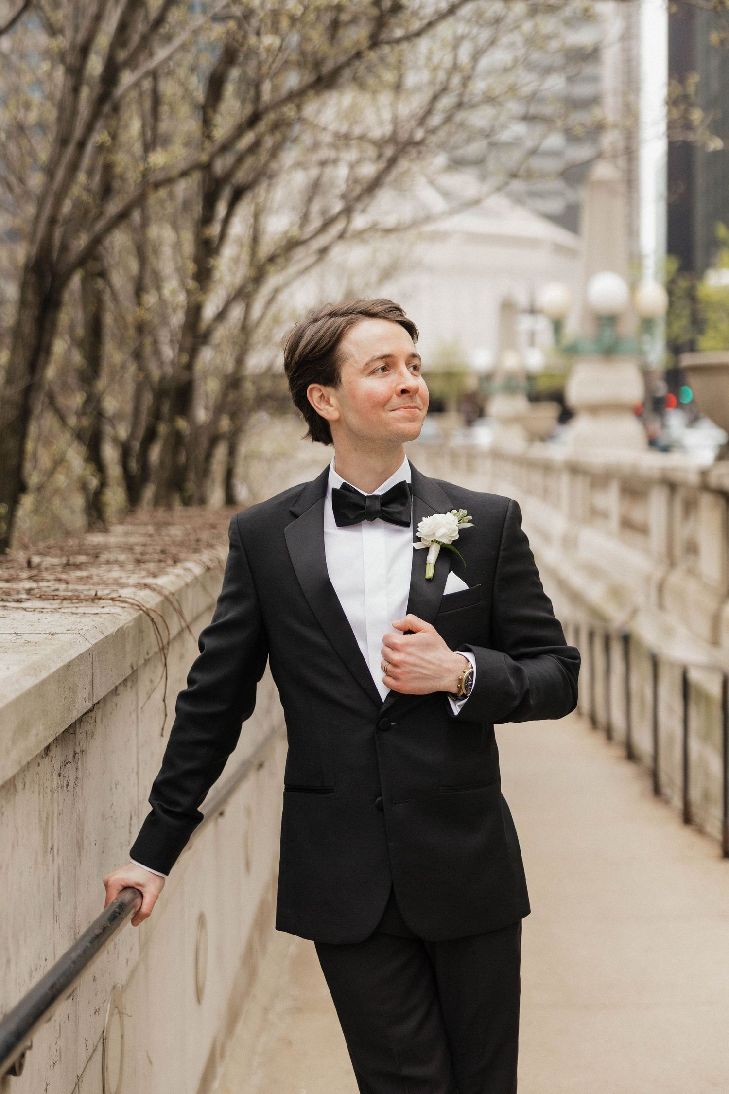 A dapper groom in a black tuxedo with bow tie and boutonniere walks confidently along an outdoor pathway.