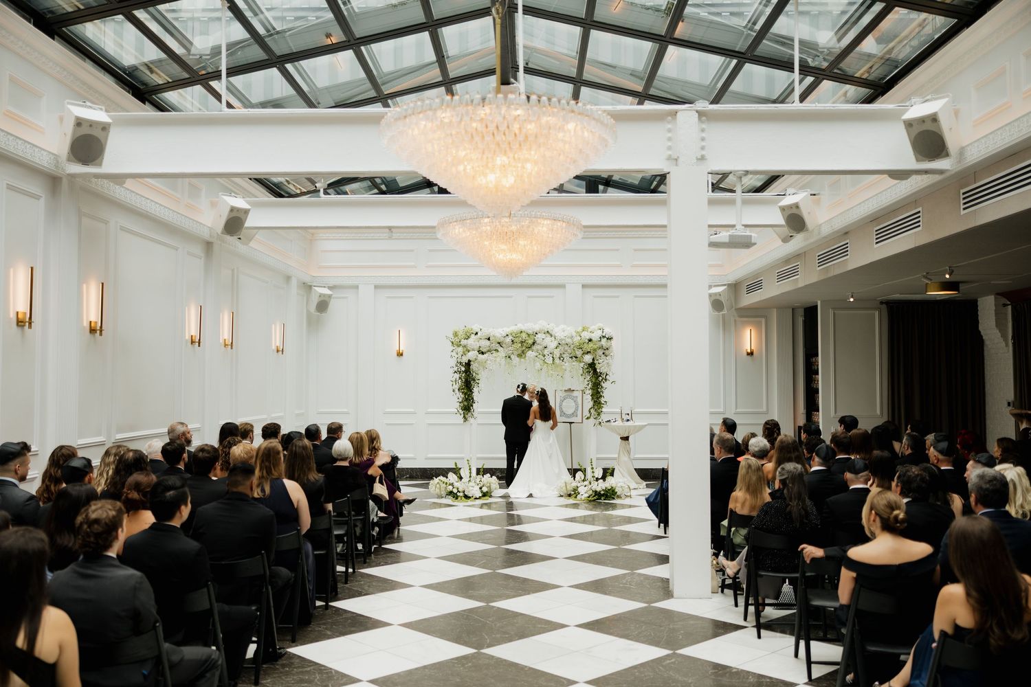 Elegant wedding ceremony in a grand ballroom with checkered floors, chandelier, and floral arch altar.