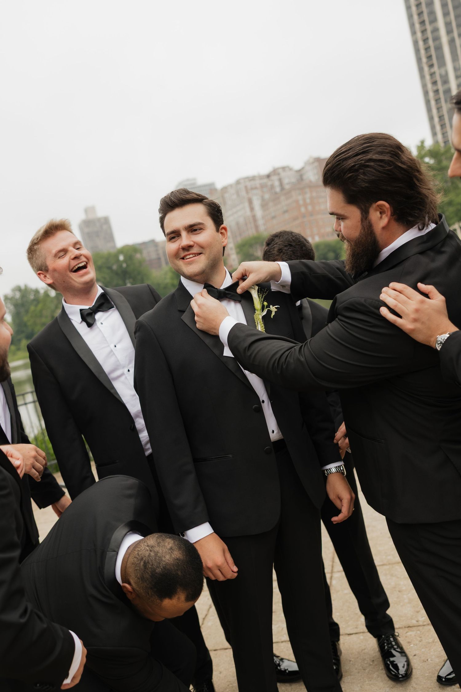 Groomsmen in black tuxedos helping groom adjust his bow tie outdoors before wedding ceremony.