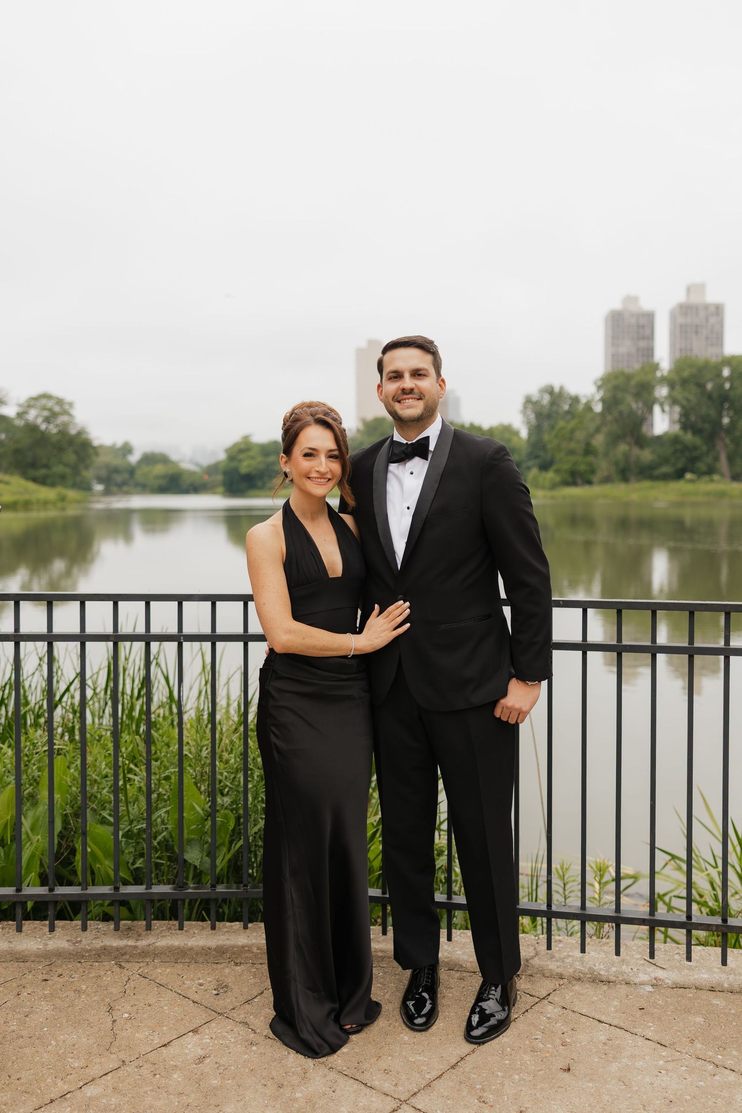 Couple in formal black attire posing by a railing with a serene lake and city skyline backdrop.