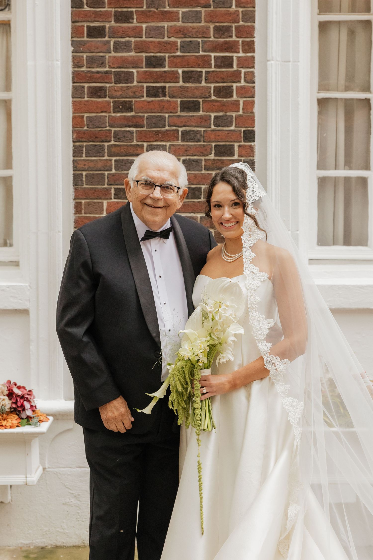 Bride in white gown and veil holds bouquet beside elderly man in tuxedo before brick building.