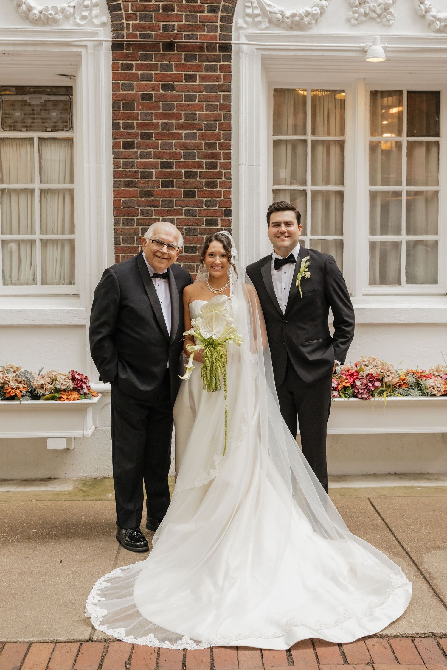 Bride in white gown with groom in tuxedo and older gentleman posing outside brick building on wedding day.