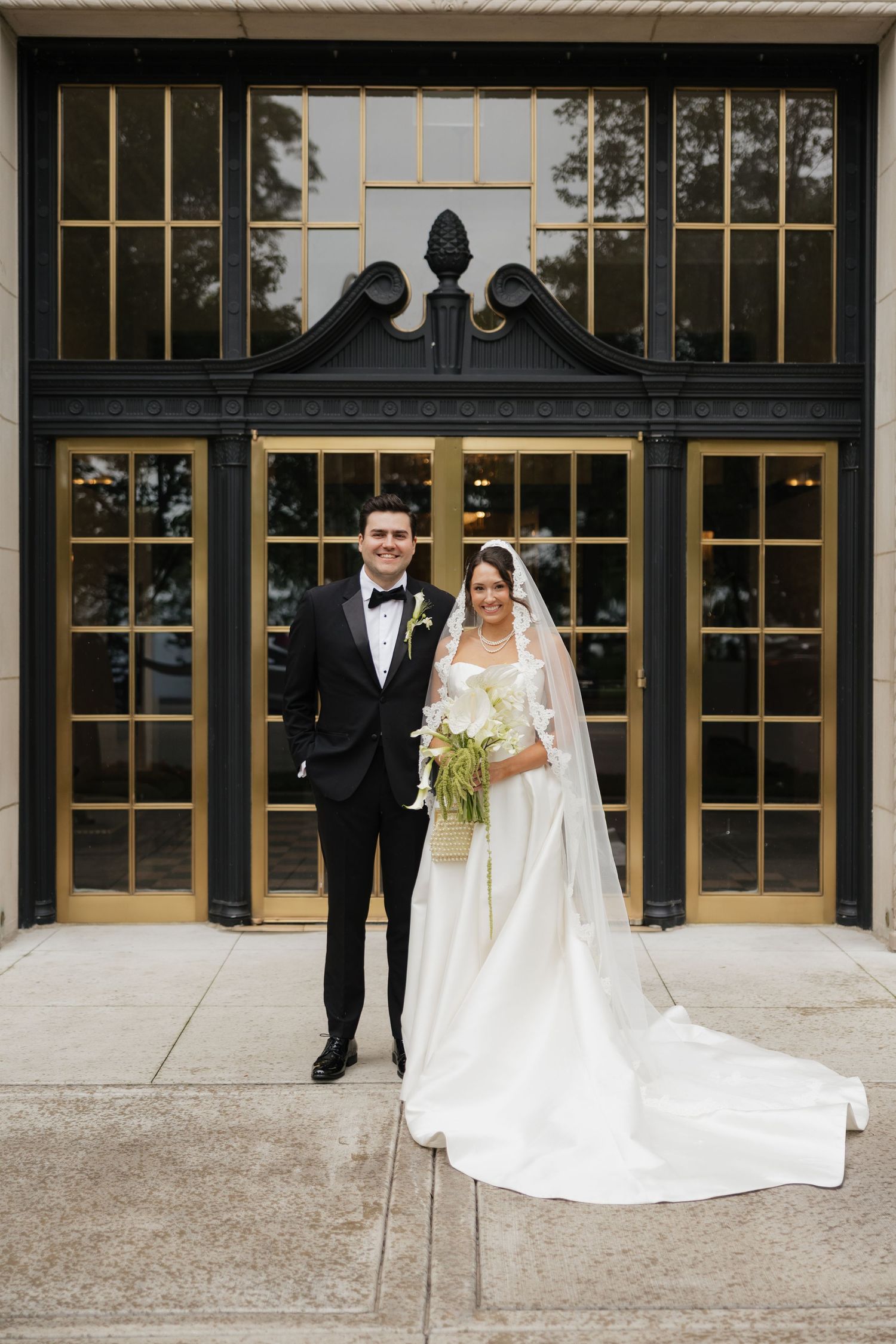 Bride and groom pose in front of elegant gold and black doors, bride wearing a long white gown with veil and bouquet.