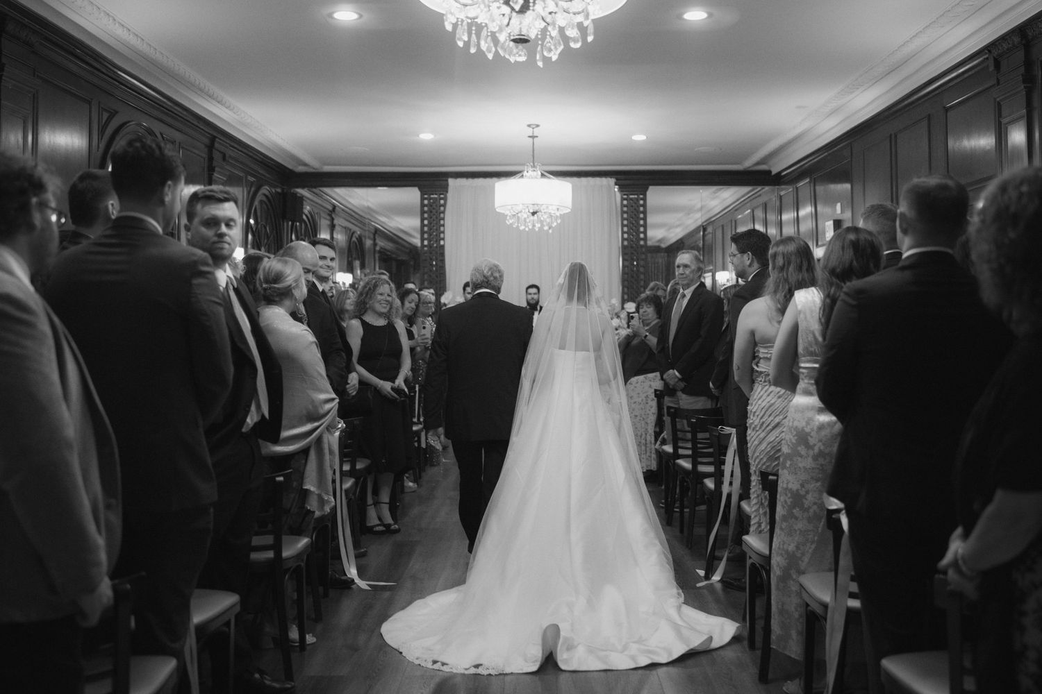 Black and white photo of a bride walking down the aisle with her father at an elegant indoor wedding ceremony.