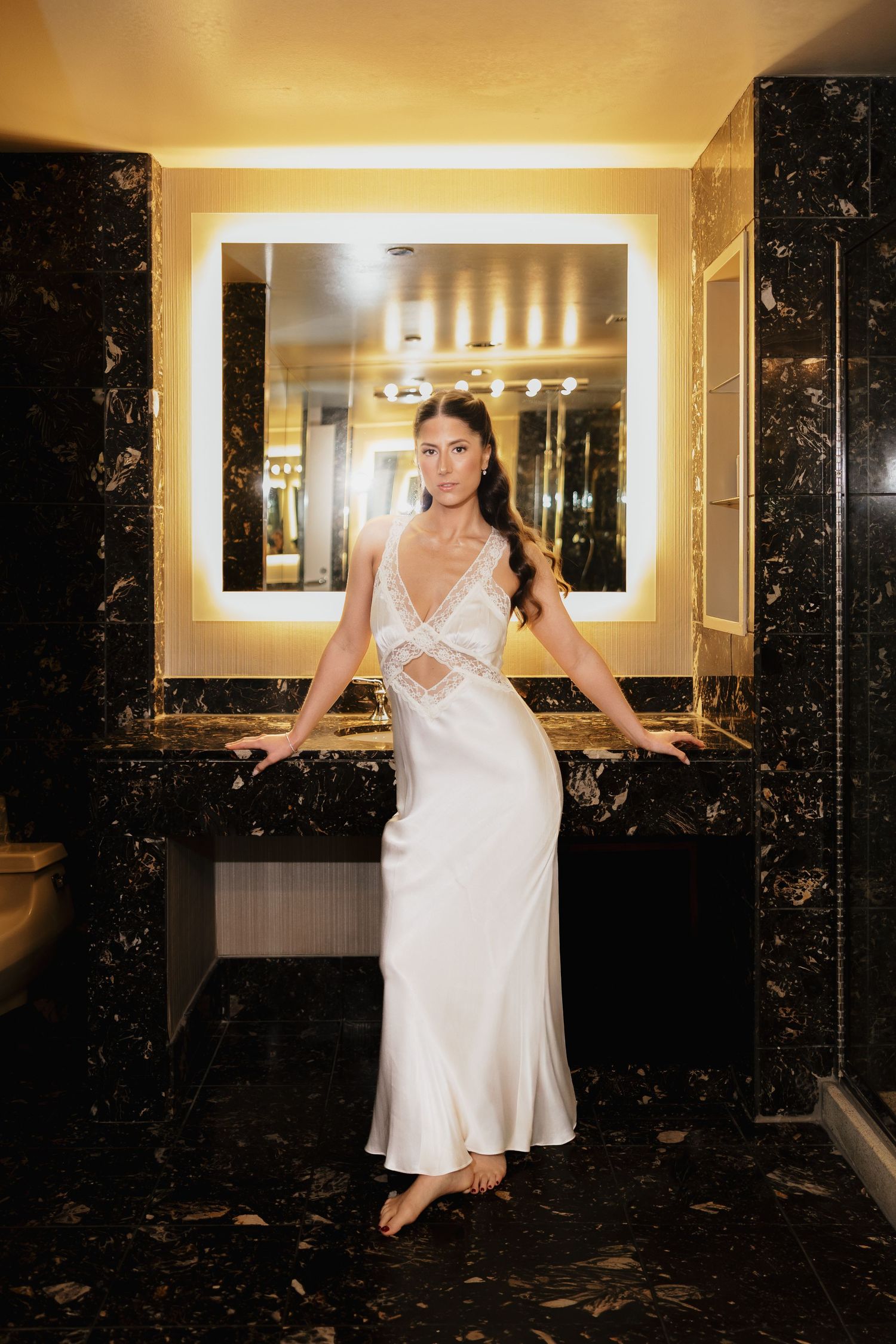 Bride poses in elegant white satin slip gown in luxurious marble bathroom with illuminated mirror.
