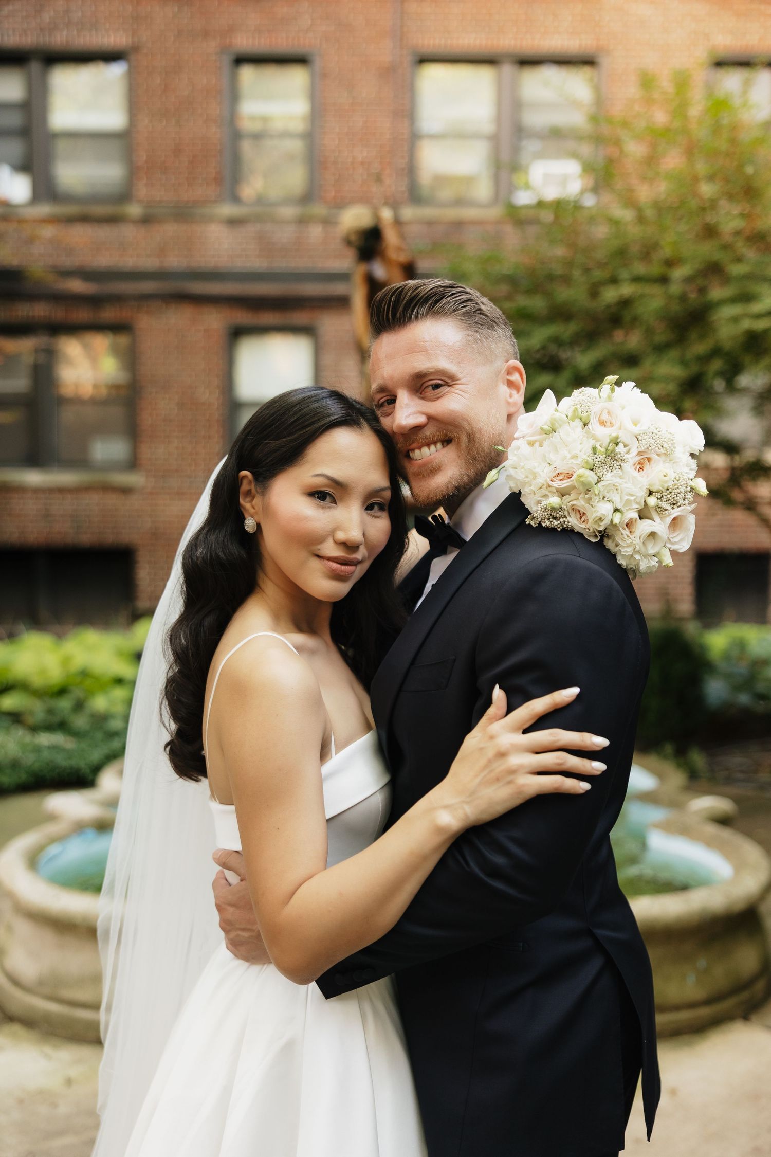 Smiling bride in a white gown and veil embraces her groom in a black tuxedo holding a white floral bouquet in a brick courtyard garden.