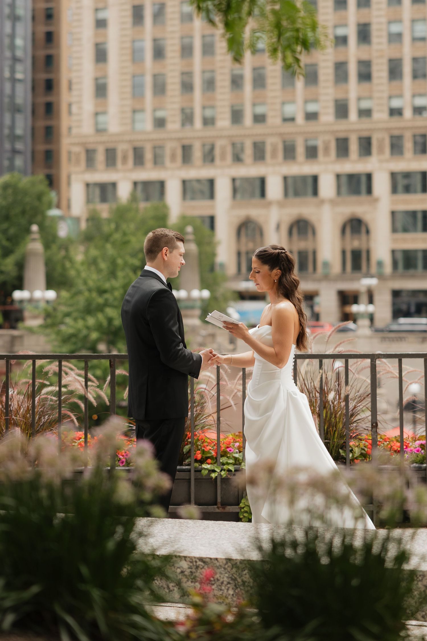 Bride in a white gown and groom in a black suit exchange vows on an urban terrace with colorful flowers and a city building behind them.