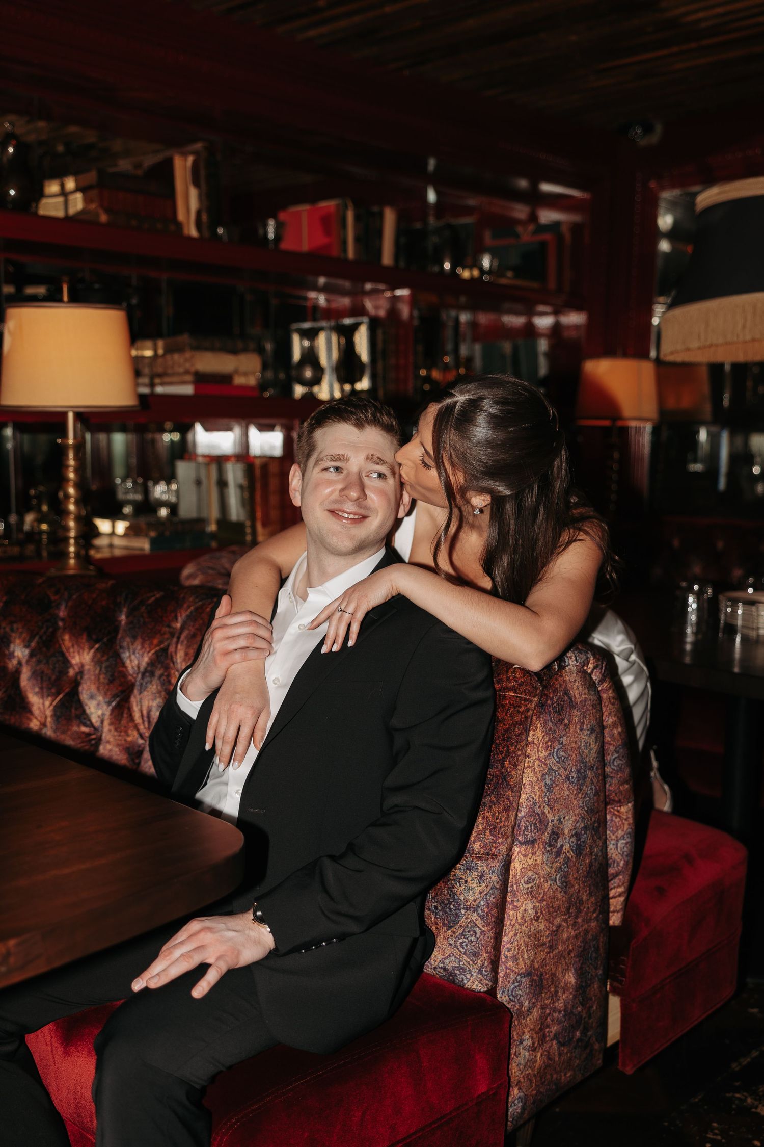 A romantic moment captured at a dimly lit bar with red leather booth seating and warm ambient lighting.