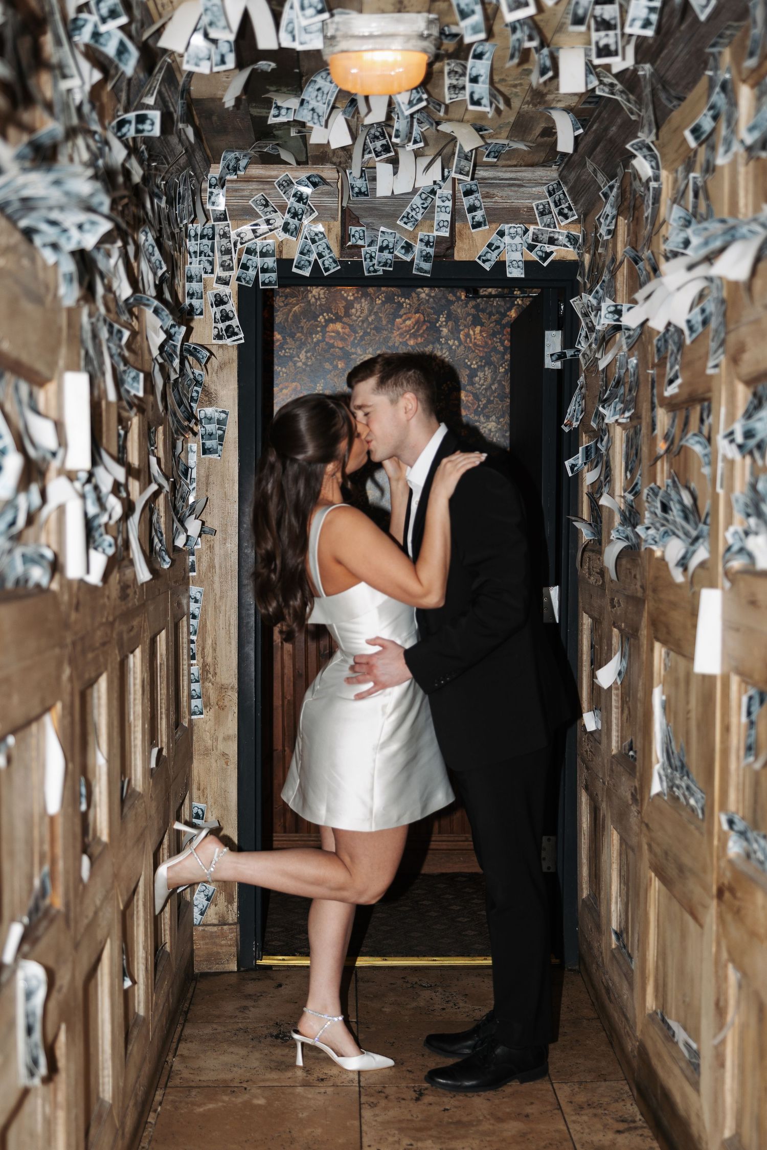 A couple shares a romantic kiss in a hallway decorated with silver streamers and warm lighting at a wedding reception.