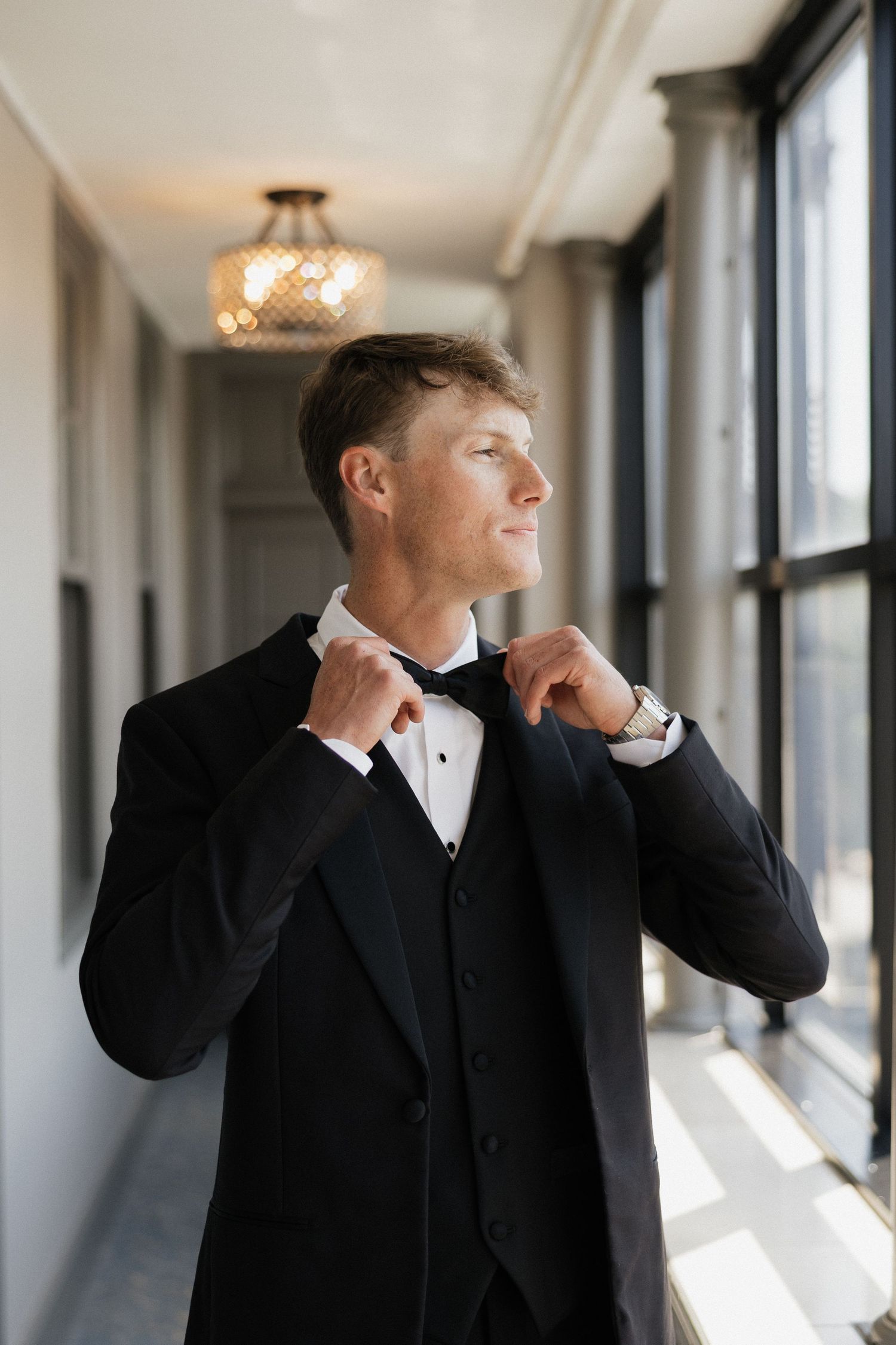 Groom in black tuxedo adjusting bow tie in elegant hallway with large windows and chandelier.