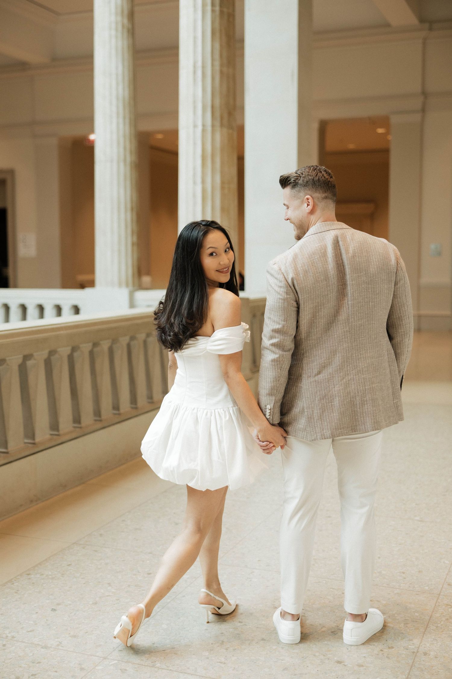Couple holding hands in elegant museum hall, woman in white off-shoulder dress looks back smiling, man in beige blazer.
