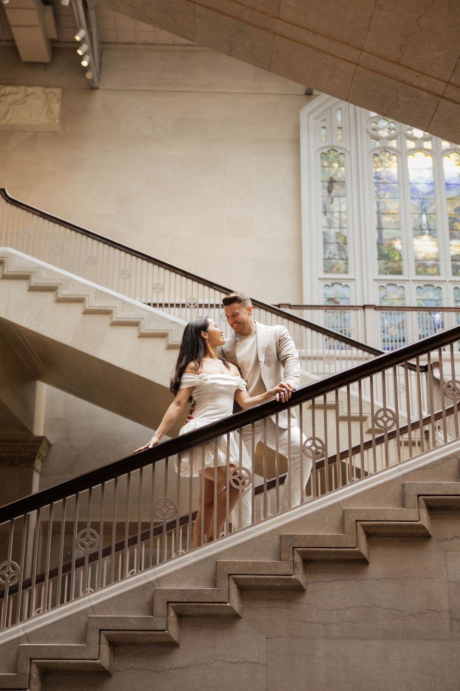 Couple in white outfits sharing a romantic moment on elegant staircase with ornate railings and stained glass window.